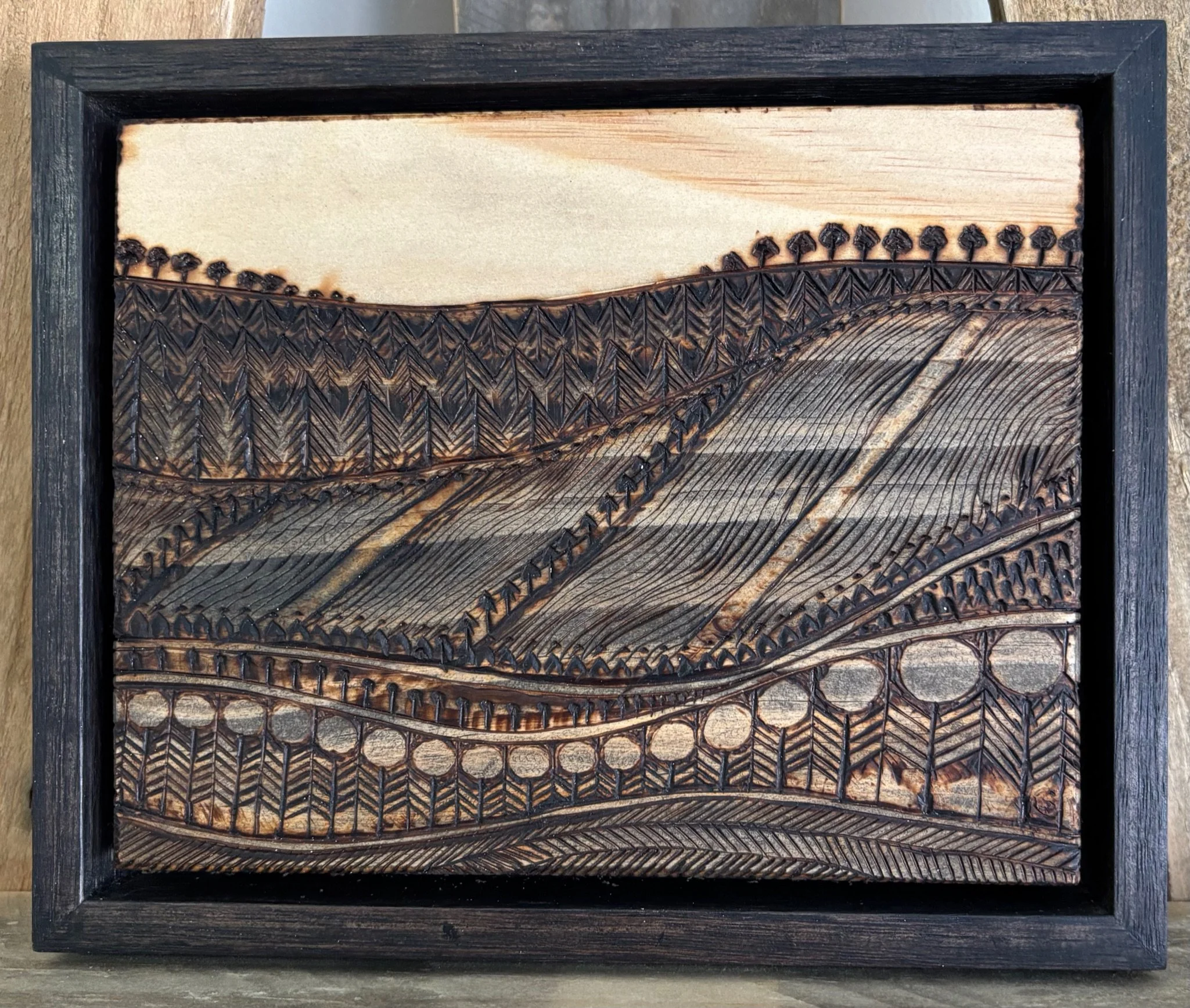 Wooden artwork depicting a landscape of hills, trees, and a bridge, carved with intricate patterns and framed in black.