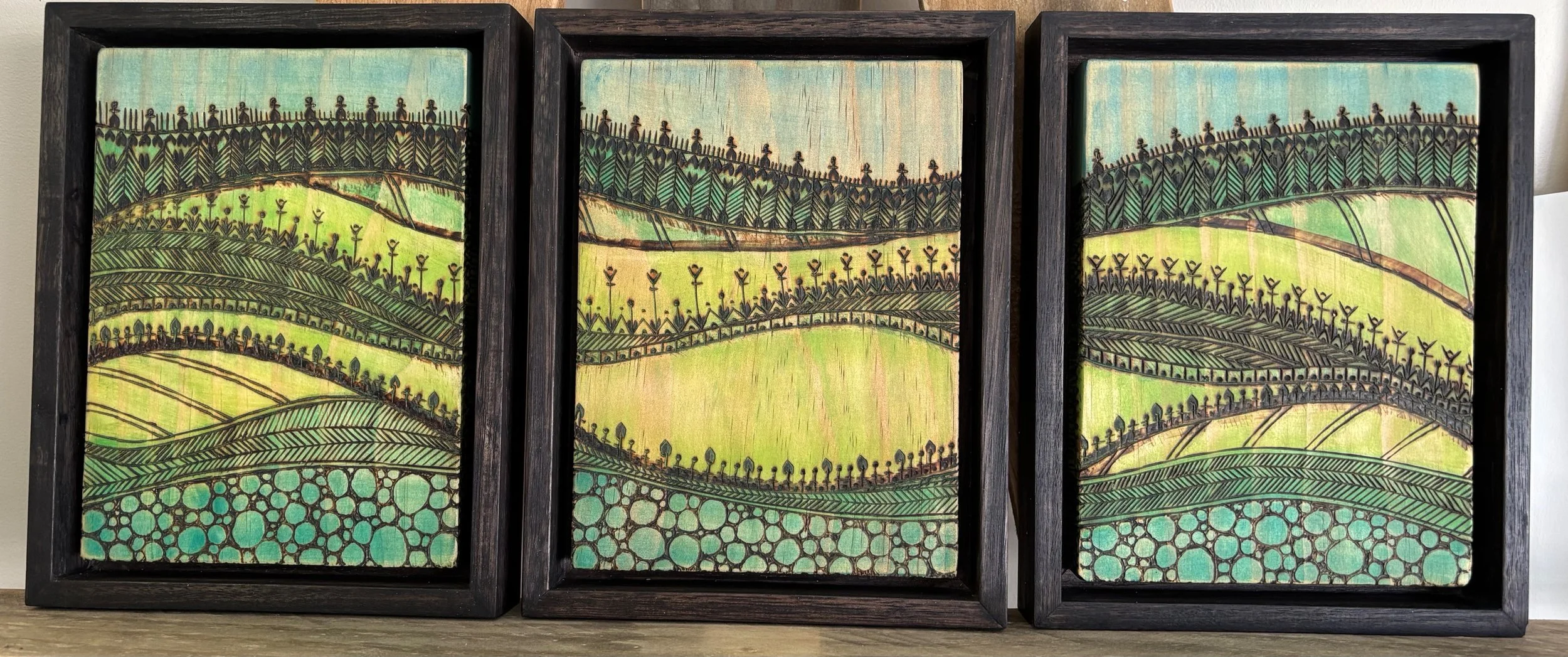 A triptych framed artwork depicting a stylized landscape of rolling hills with patterned vegetation and a pebble-like foreground in a folk art style.