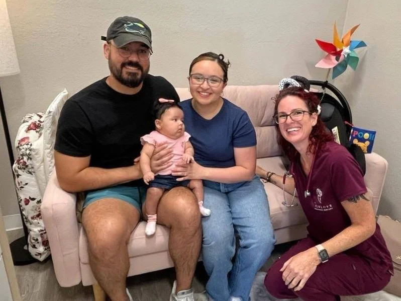 Four people, including a man, woman, girl, and baby, sitting on a pink couch in a living room, smiling and posing for a photo. The man is wearing a black cap and shirt, the woman has glasses and a navy shirt, the girl has glasses and is wearing blue jeans, and the baby is dressed in pink and white.