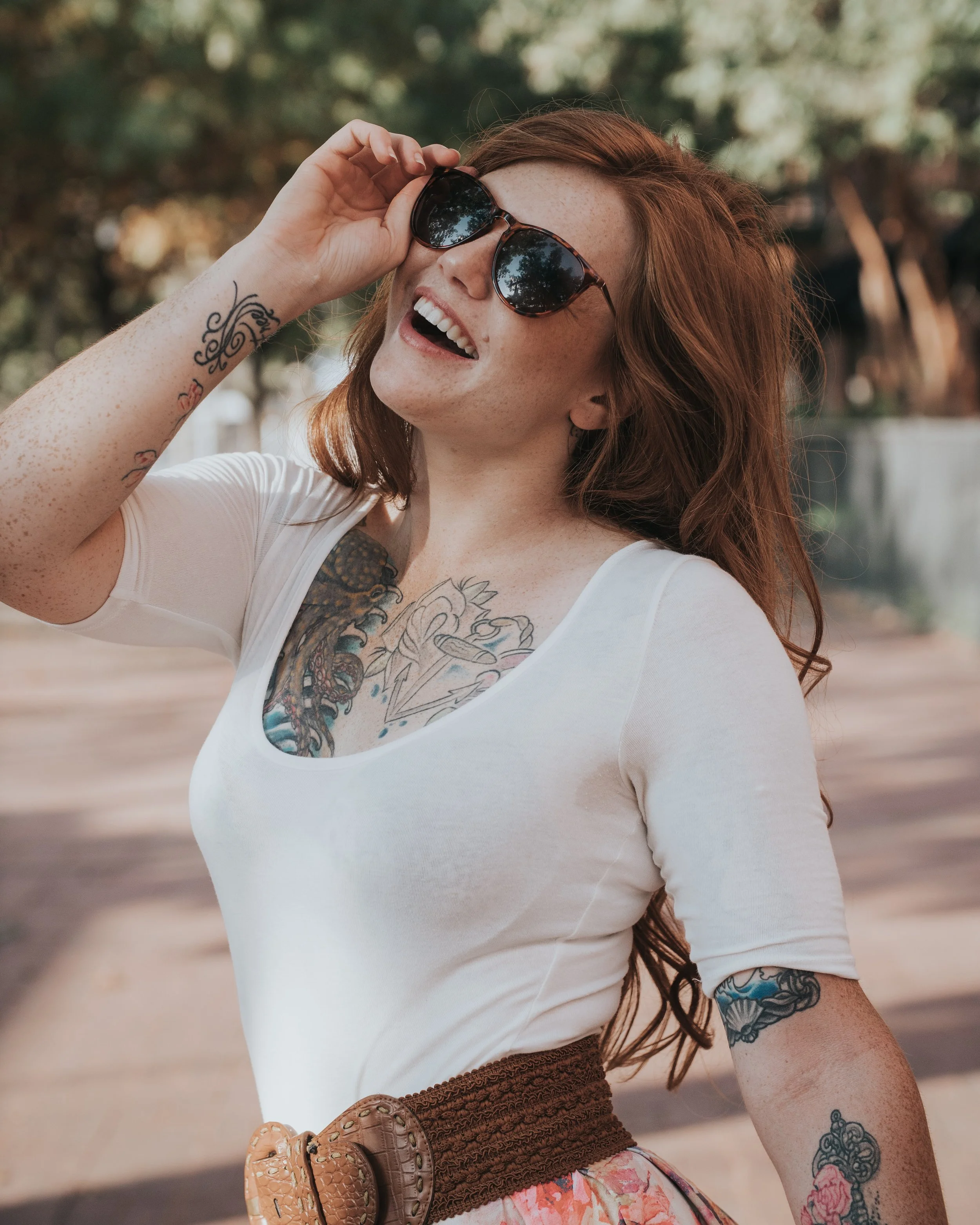 A smiling woman with red hair and sunglasses outdoors, wearing a white shirt, tattoos, and a brown belt with a large buckle.