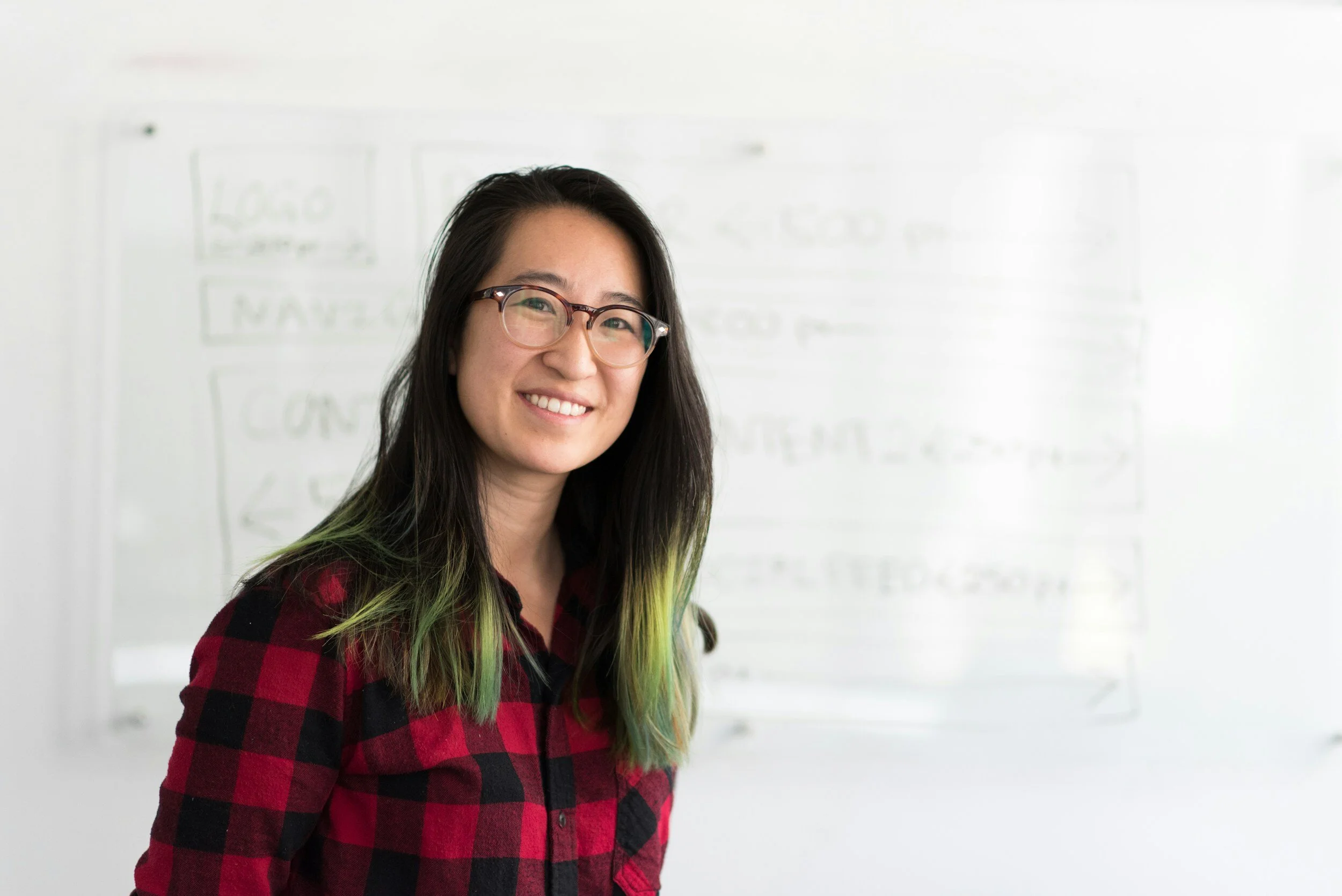 A woman with long black hair with green tips, wearing glasses and a red and black checkered shirt, smiling in front of a whiteboard with writing.
