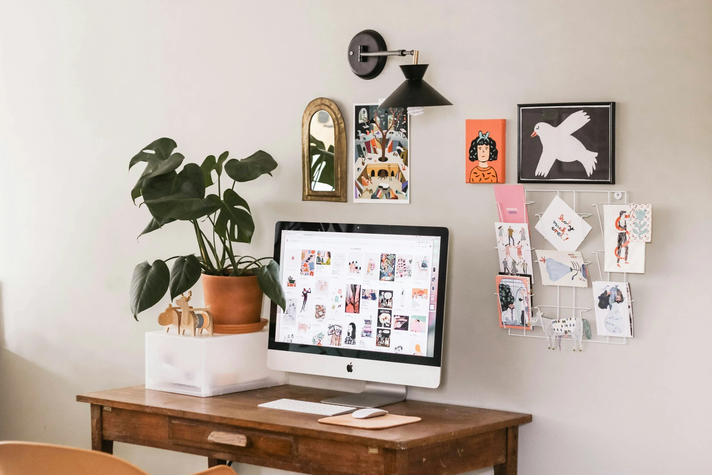 A home workspace with a wooden desk, Apple iMac computer, potted plant, decorative wall art, and a memo board with postcards and cards.