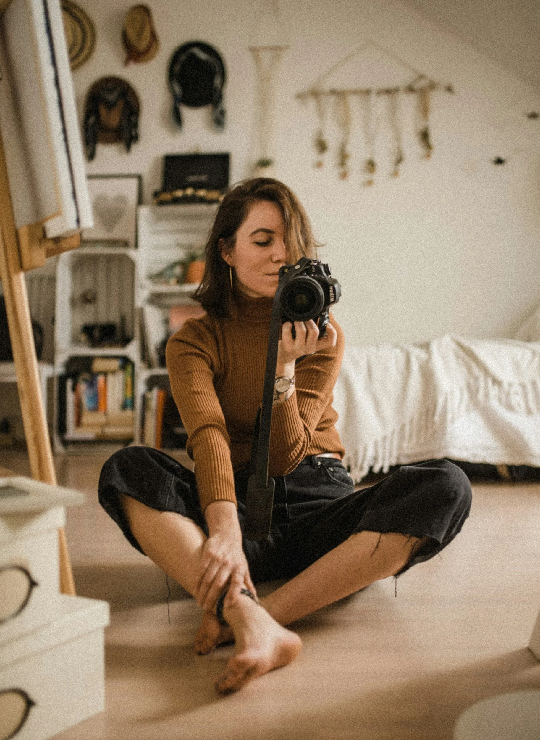 A woman sitting on the floor, taking a selfie in a mirror with a DSLR camera, in a cozy, bohemian-style bedroom decorated with wall hangings, hats, and books.