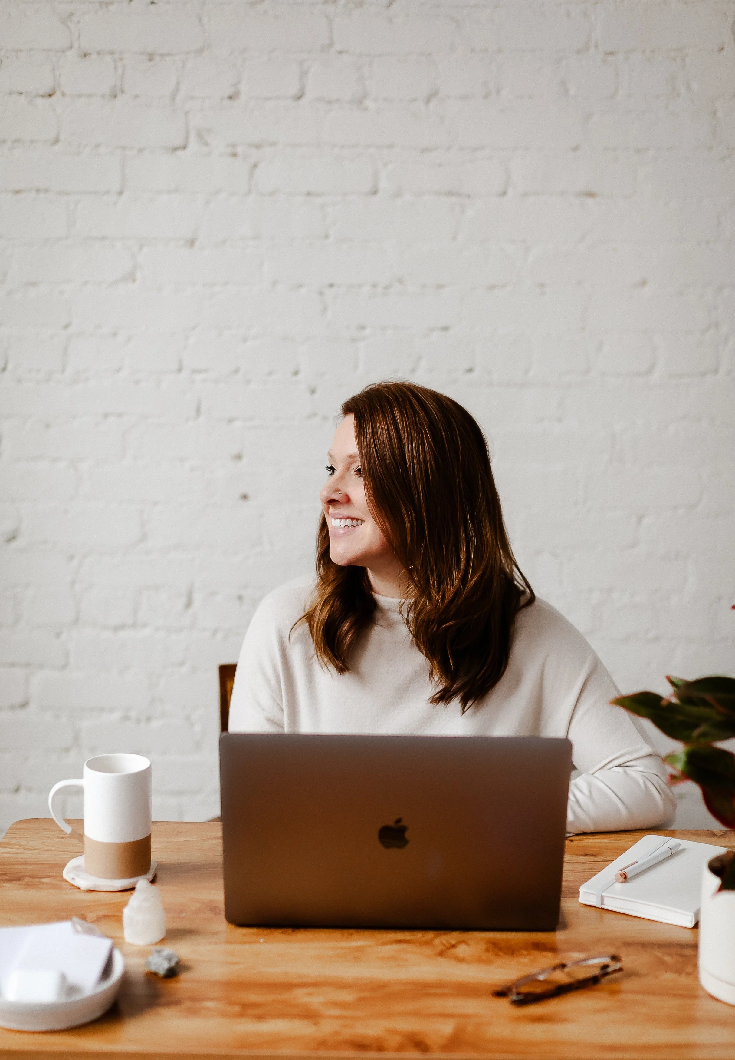A woman with long brown hair smiles while sitting at a wooden desk with a silver laptop, a white mug, a notebook with a pen, a pair of glasses, and some decorative items, against a white brick wall.