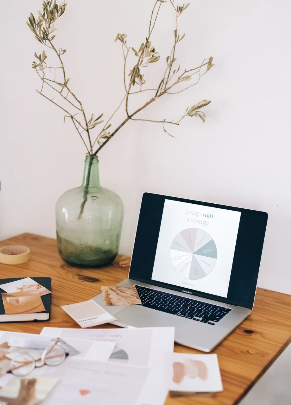 A wooden desk holding scattered papers, eyeglasses, a black notebook, and a MacBook laptop displaying a pie chart, with a green glass vase containing dried branches against a plain wall.