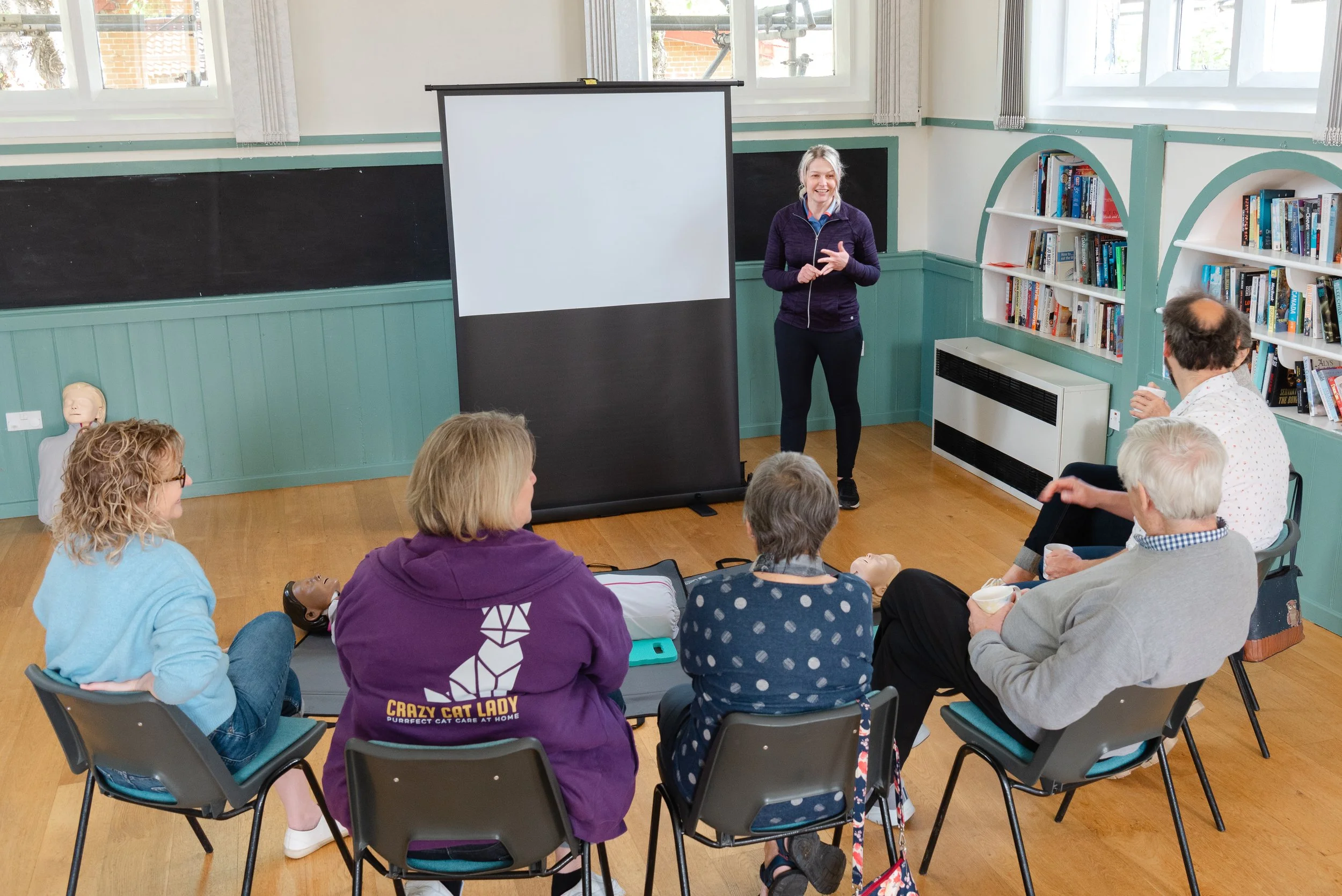 An image showing a training room.  The instructor is stood by a pull up display screen.  They are facing a semi circle of people sat in chairs.