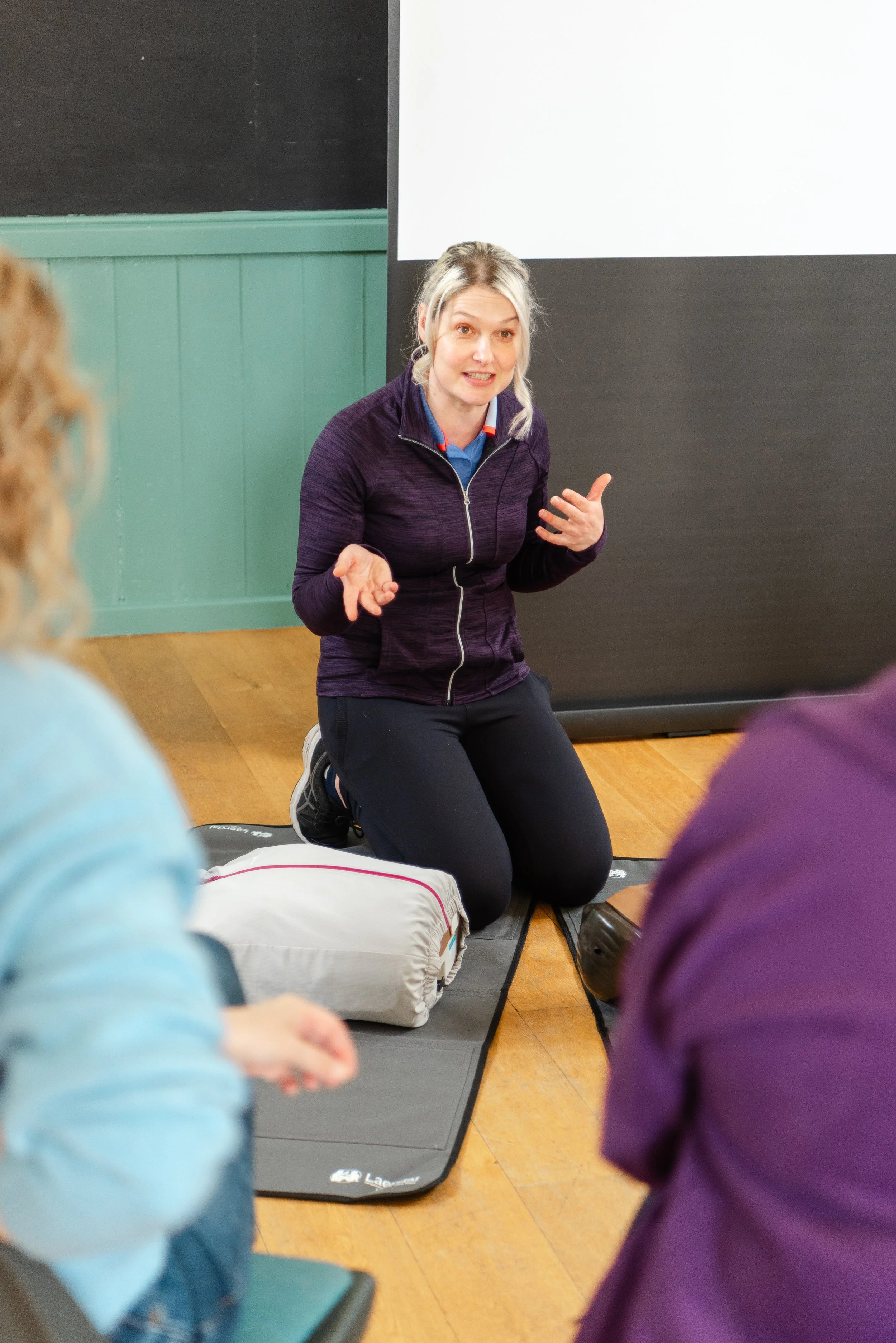 An image showing a first aid instructor.  They are wearning back trousers and a purple zip up jacket.  They are knelt down and discussing training with a group of learners