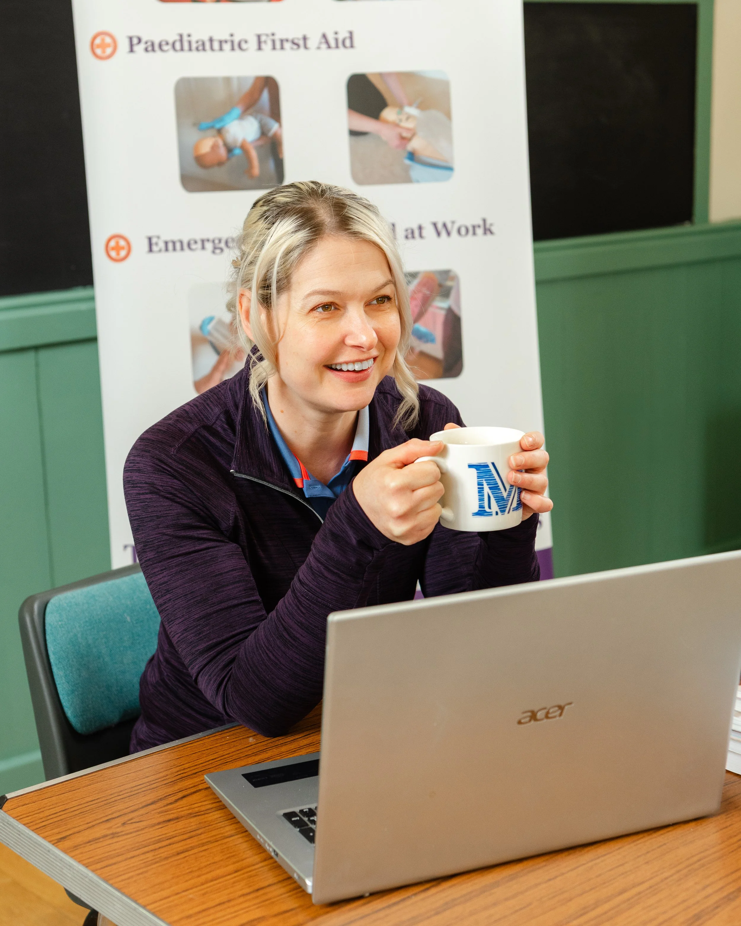 A woman with blonde hair smiling and holding a mug sitting at a desk with a laptop. In the background, there is a poster with images related to pediatric first aid and emergency response.