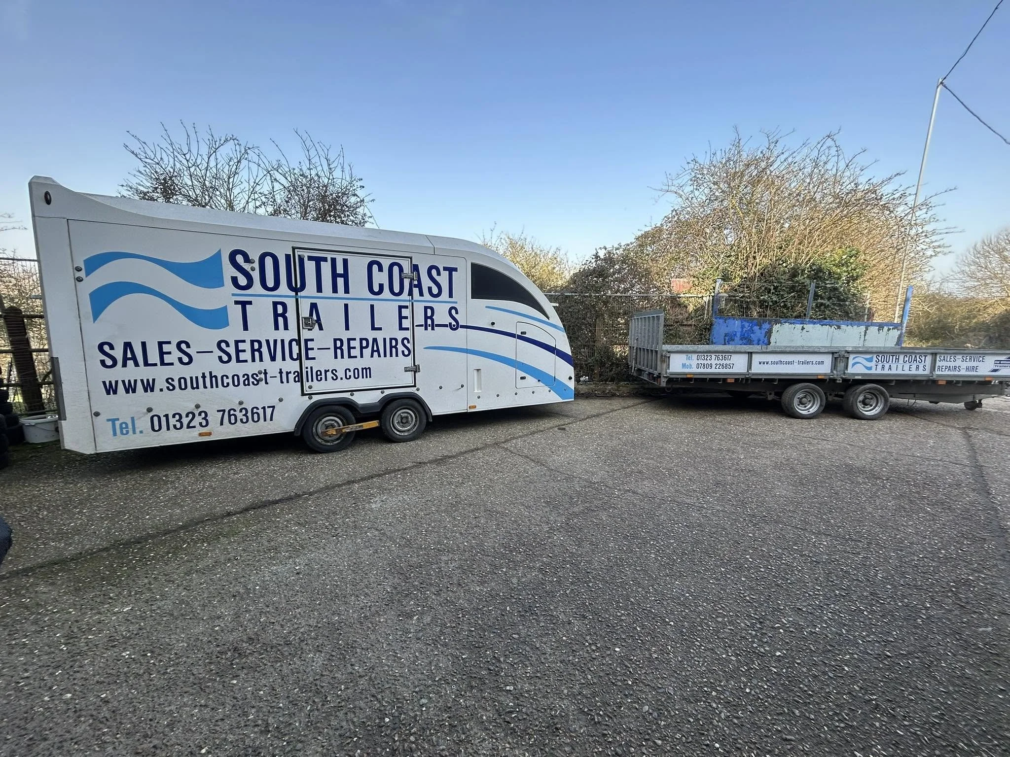 A white trailer with blue text advertising South Coast Trailers, parked on a gravel lot with trees and a clear blue sky in the background.