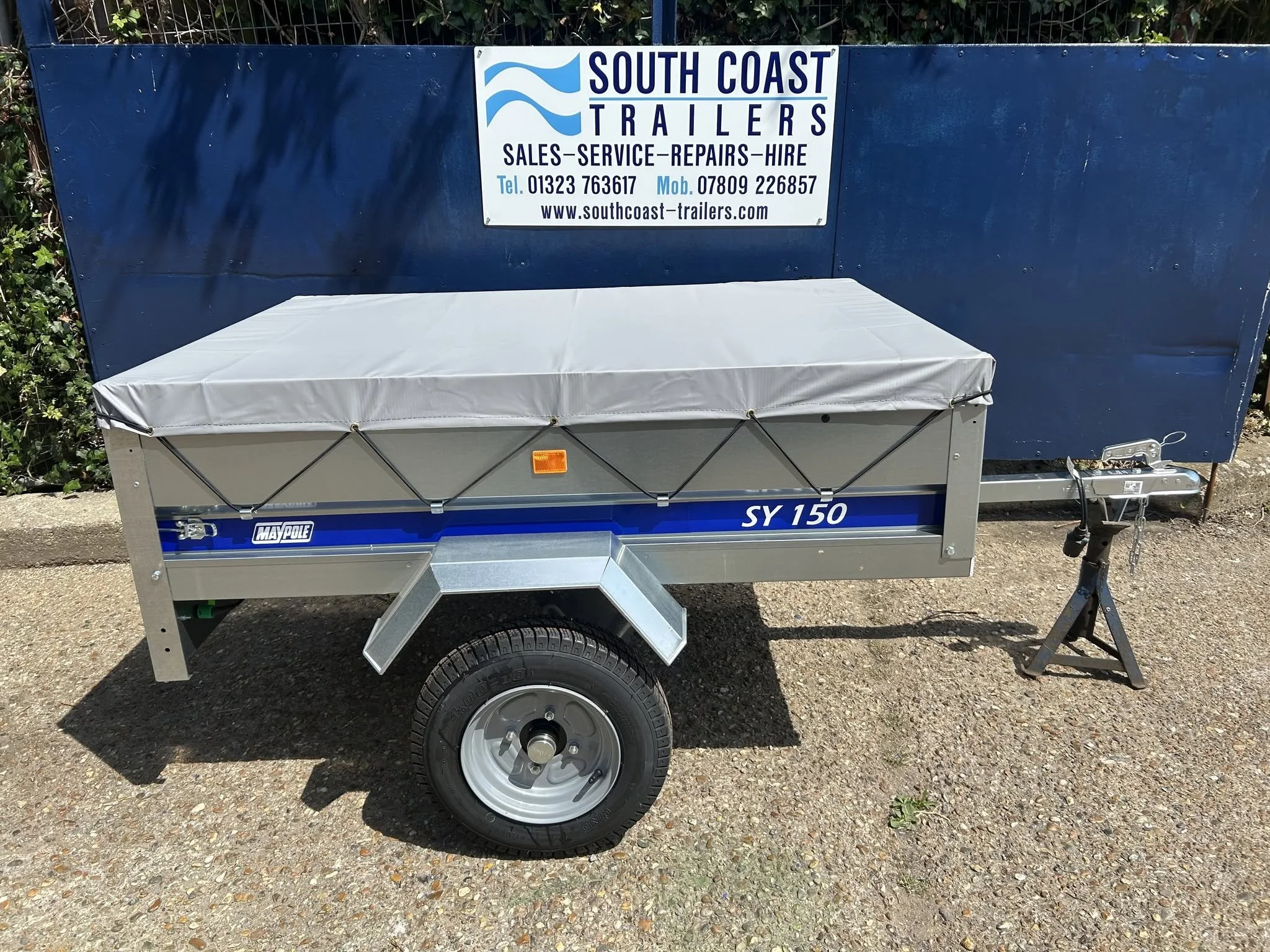 A small trailer with a blue cover and a sign for South Coast Trailers, displaying contact information and services, parked on a gravel surface with greenery behind.