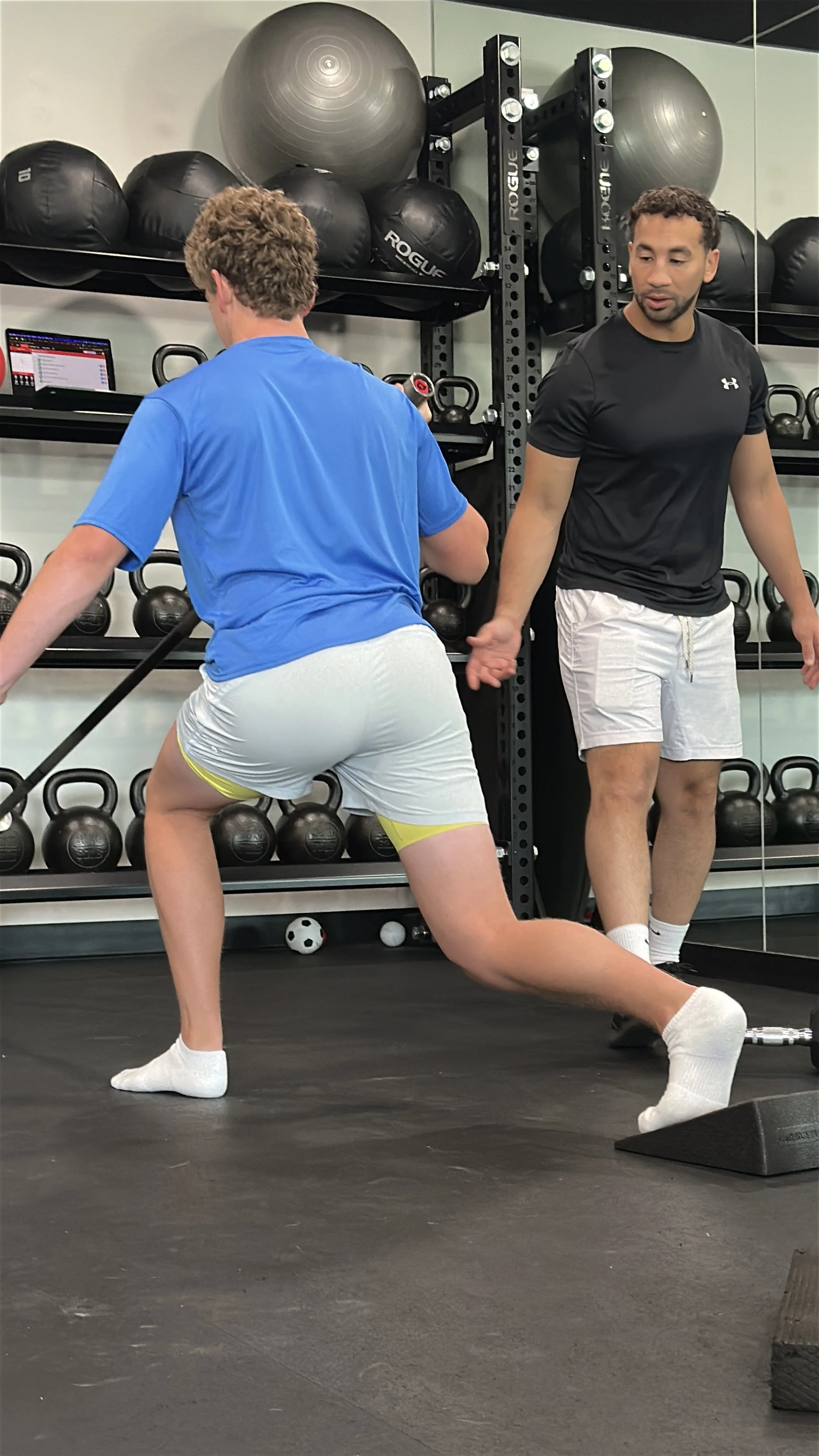 A man in a black athletic shirt and white shorts assisting a man in a blue t-shirt and white shorts during a workout at a gym. The man being assisted is performing a knee bend with a resistance band around his thighs, on a black rubber gym floor. Gyms equipment, including medicine balls and kettlebells, are visible in the background.