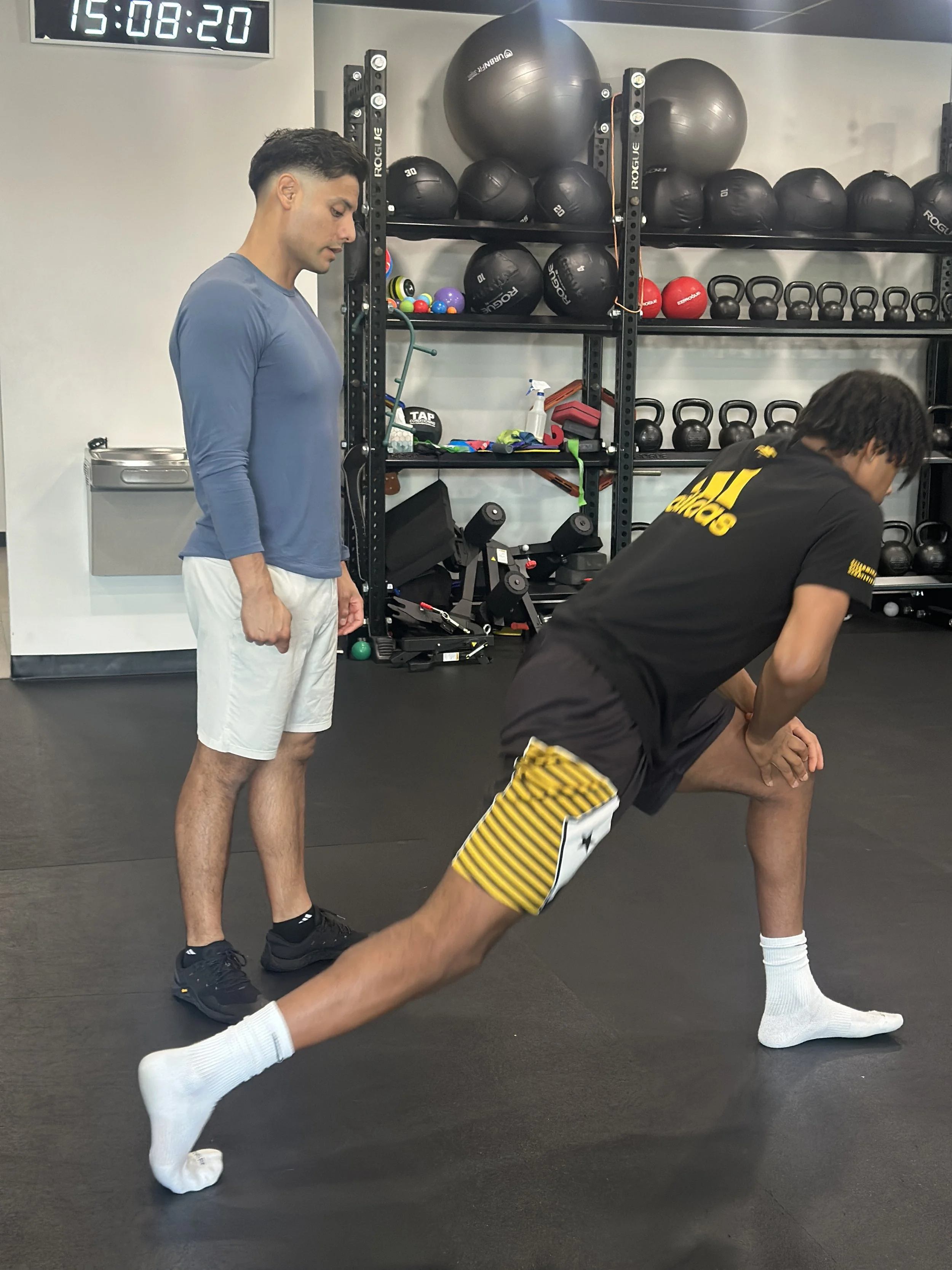 A person is performing a stretching exercise with one leg extended behind, and two individuals, possibly trainers, are observing him in a gym with various fitness equipment and a digital clock showing 15:08:20.