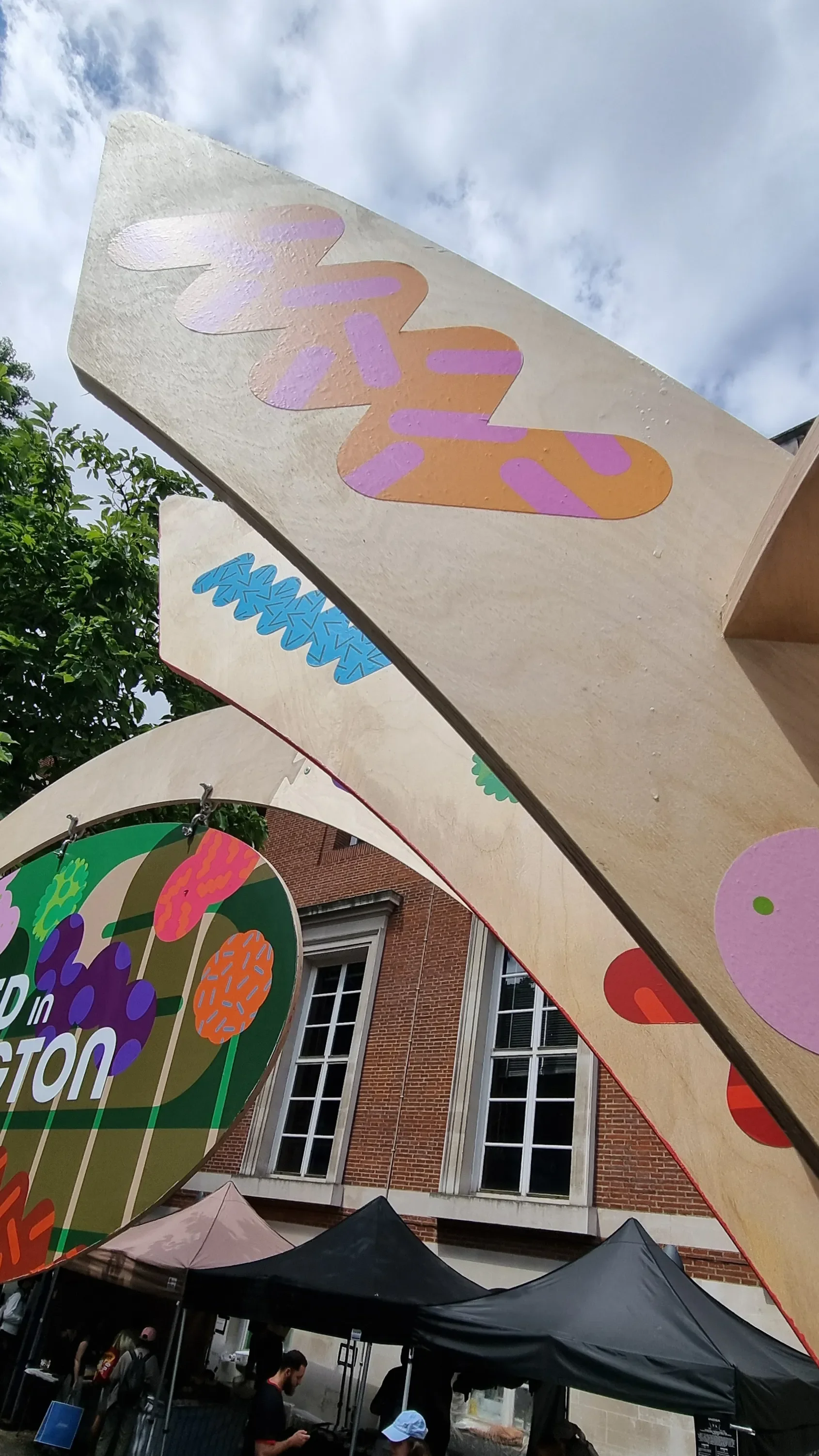 Colorful banners with abstract flower and design patterns hanging outside a building with brick walls and tall windows, indicating an outdoor market or festival.