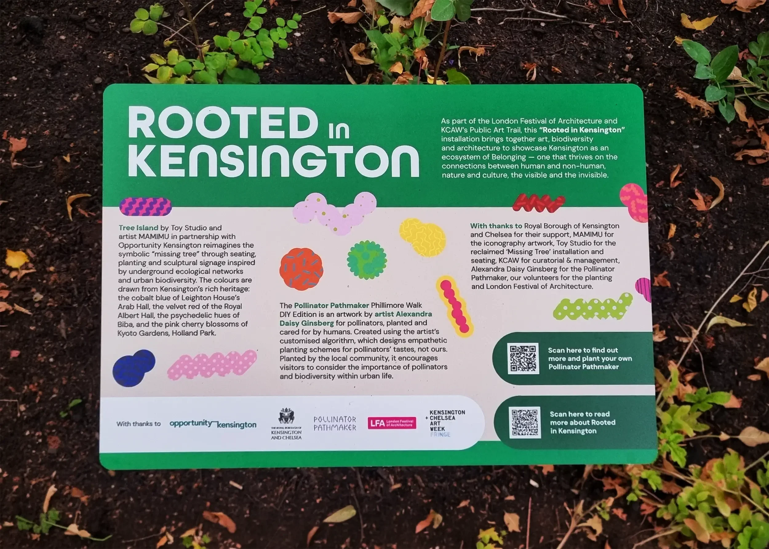 A colorful informational sign titled 'Rooted in Kensington' placed in soil among small green plants and fallen leaves. The sign contains text about a public art trail and various graphic illustrations of stylized plants and natural elements.
