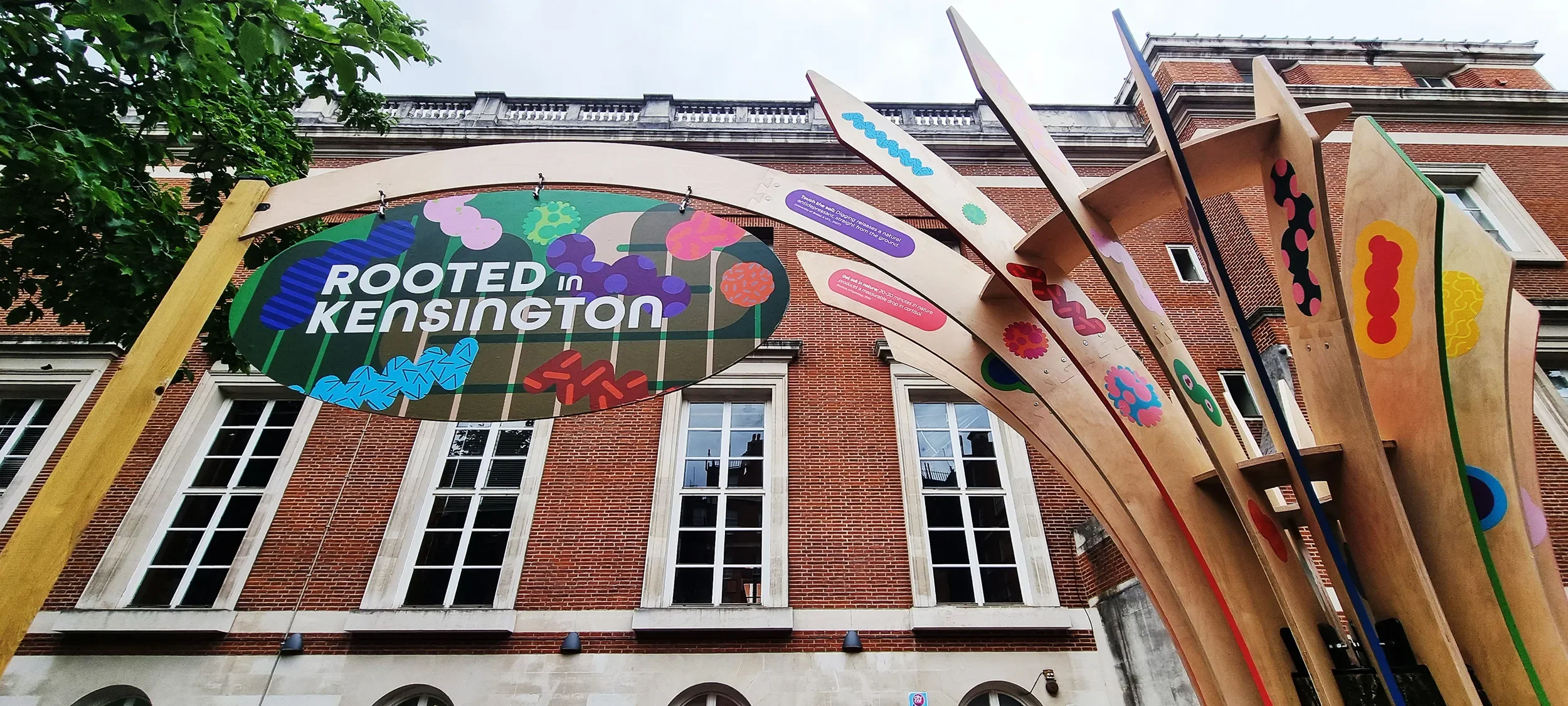 Colorful outdoor art installation featuring a sign that says 'ROOTED IN KENSINGTON' hanging from a wooden arch, with abstract, brightly colored shapes on vertical wooden panels against a brick building background.