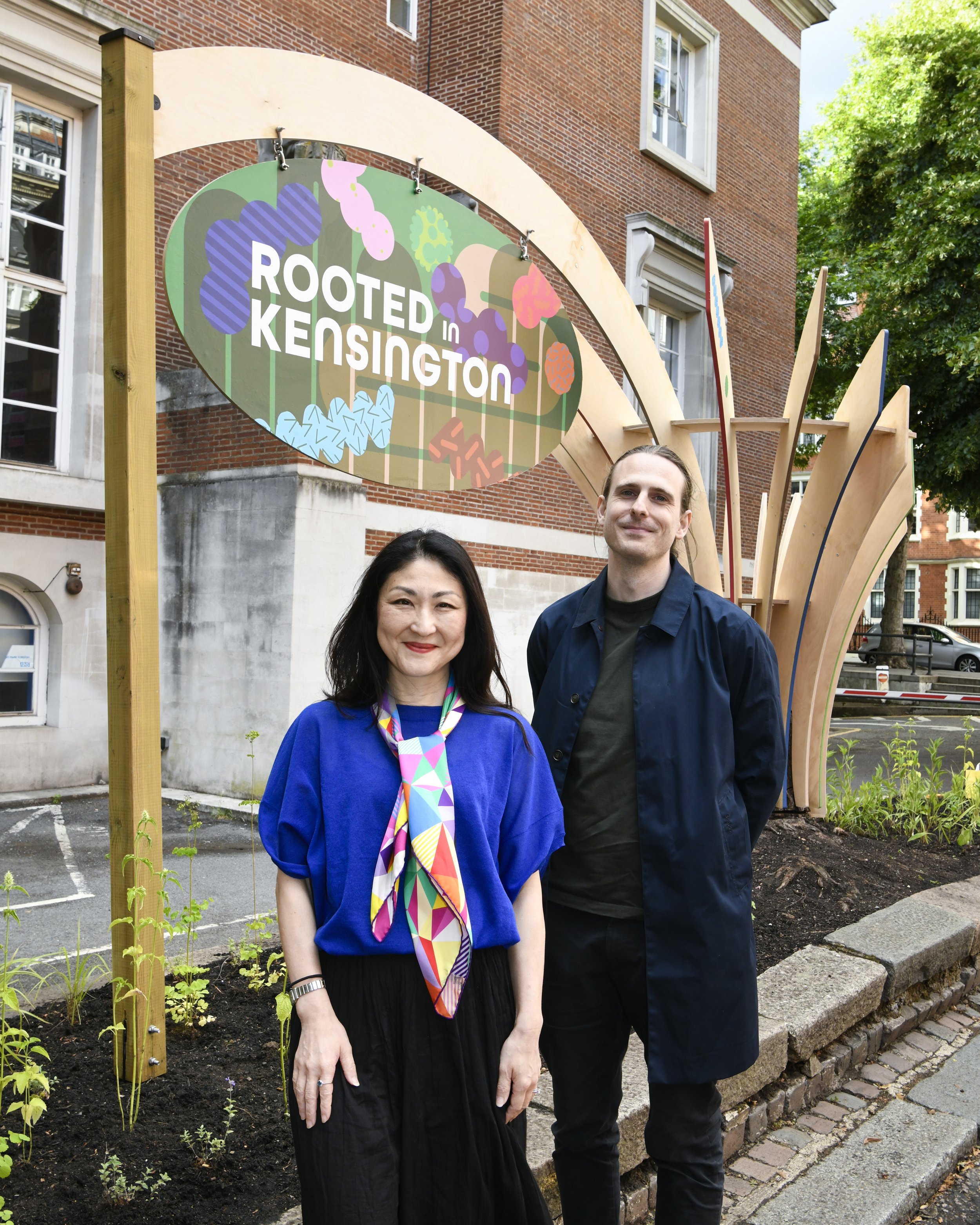 Artists MAMIMU (June Mineyama-Smithson) and Toy Studio (Toby Plunkett) standing outdoors in front of a sign that reads 'Rooted in Kensington.'