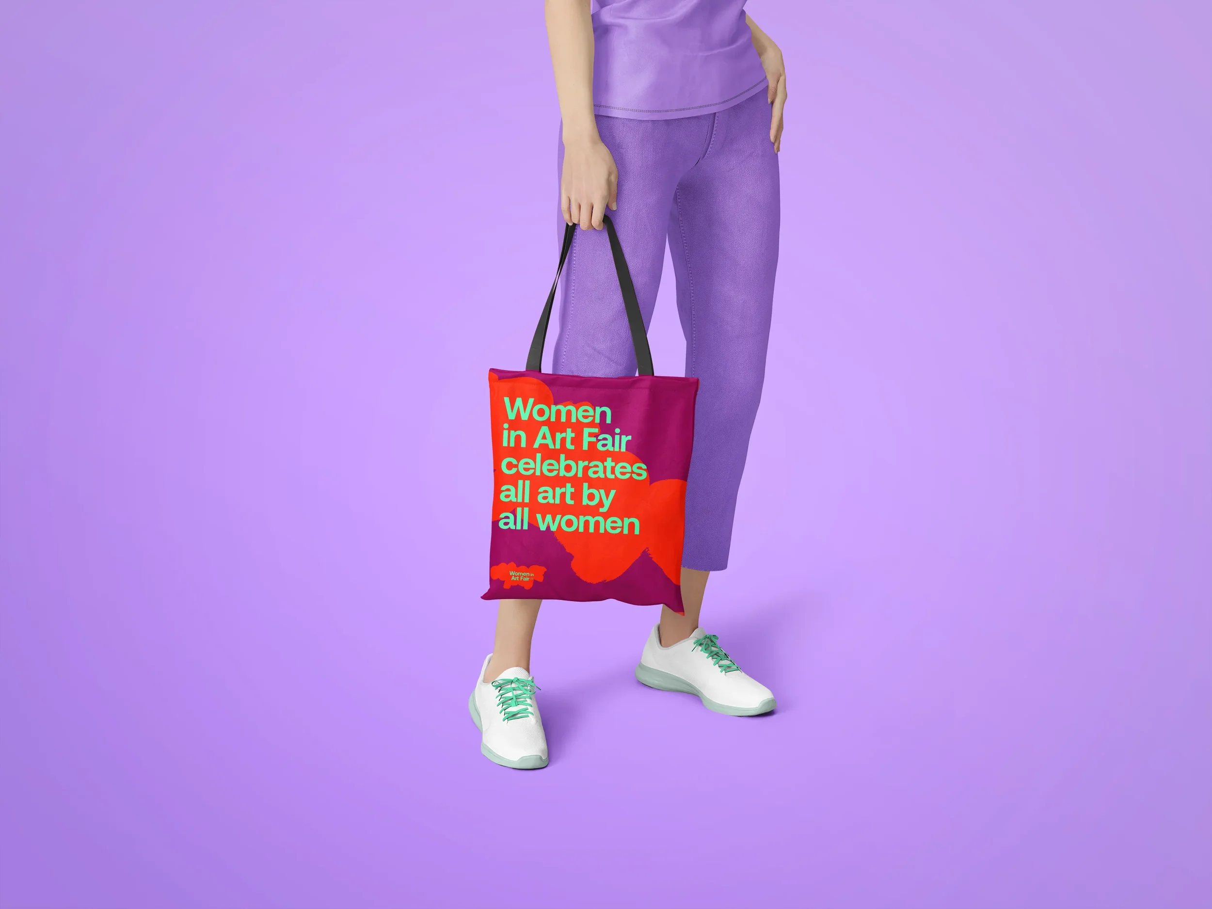 A woman holding a tote bag with colorful text, standing against a purple background. The bag reads 'Women in Art Fair celebrates all art by all women'. The person is wearing purple pants and white shoes with teal laces.
