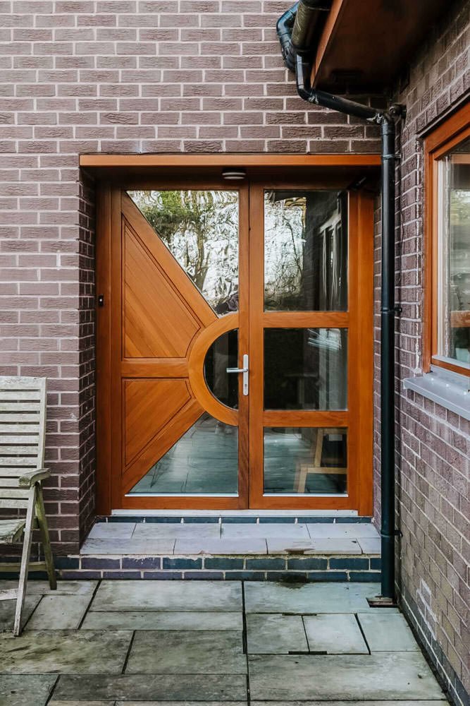 Modern wooden front door with geometric design, surrounded by brick wall and adjacent patio area.