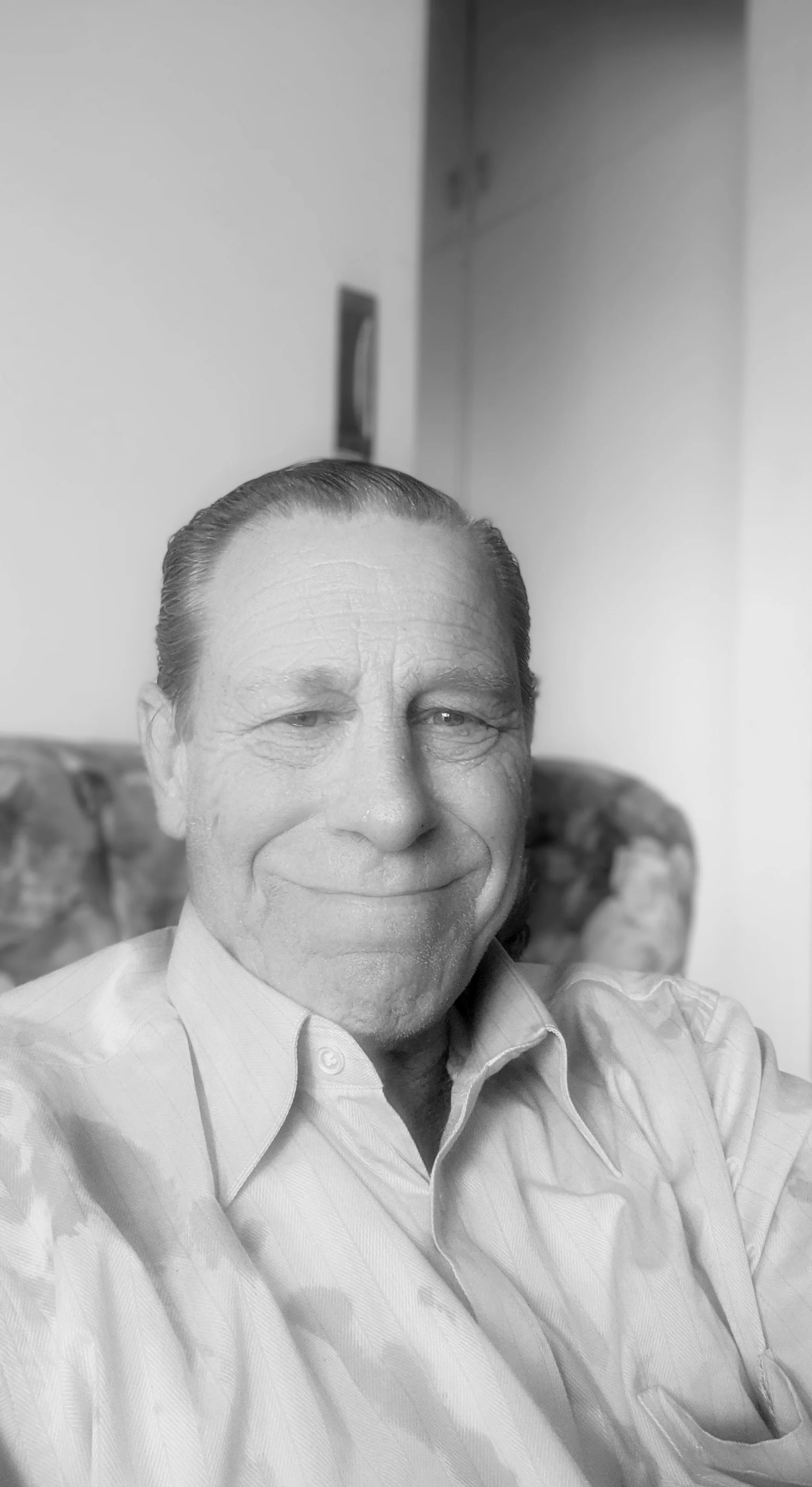 A black and white photo of a smiling man with combed-back hair, wearing a dress shirt, sitting on a floral-patterned armchair.