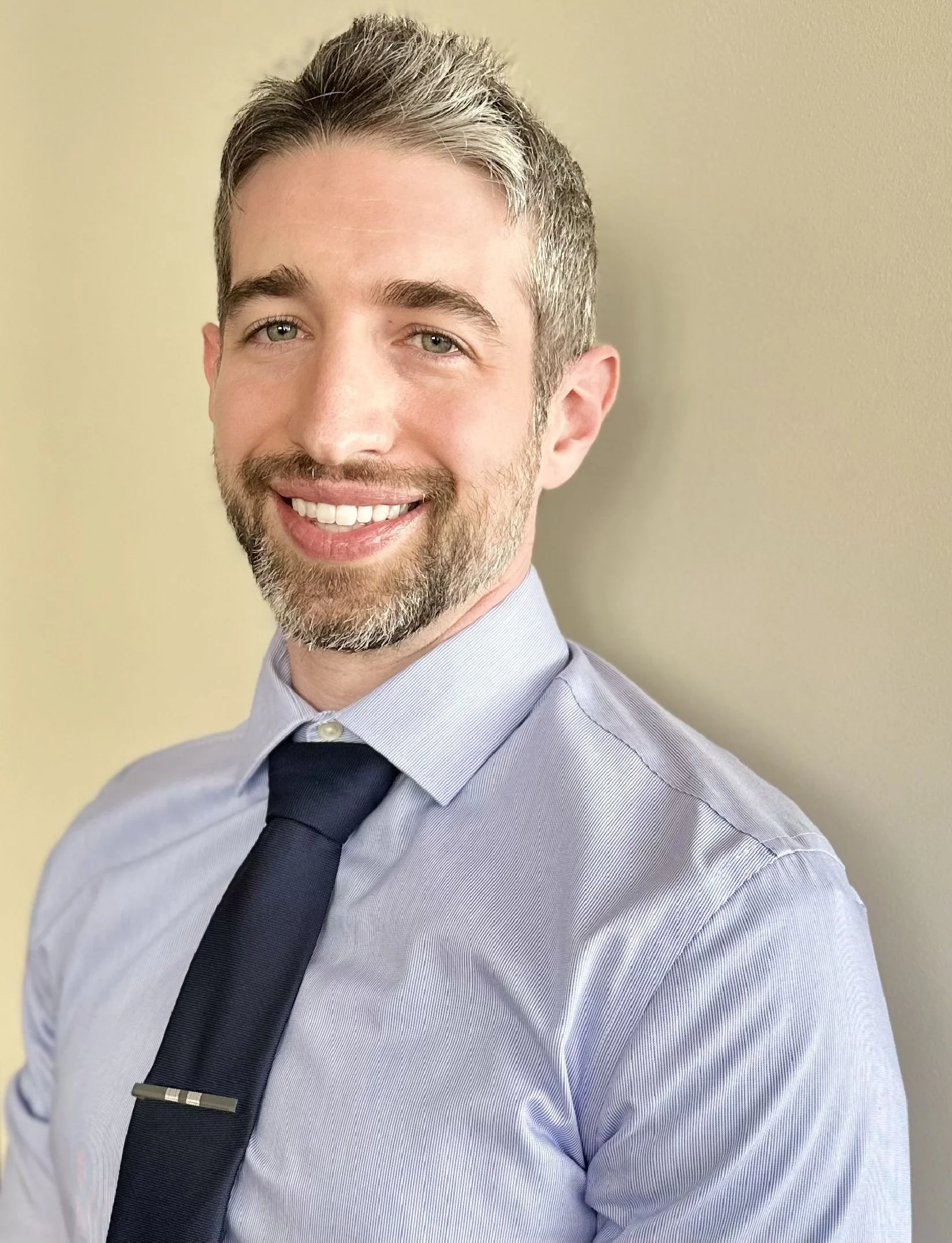 A smiling man with light brown hair and a beard, wearing a light blue dress shirt and a dark tie, standing against a beige wall.