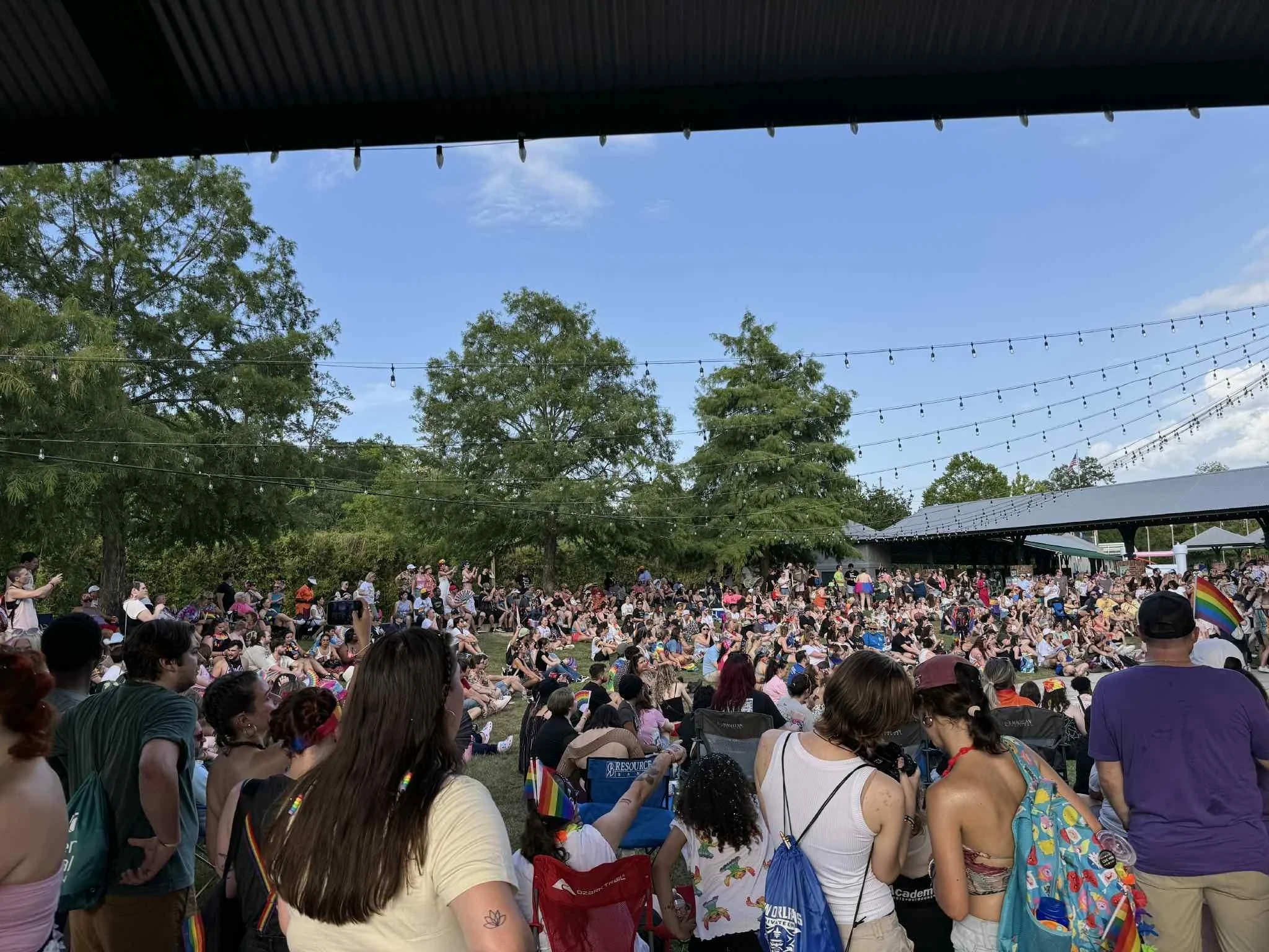 Large crowd gathered outdoors for a Pride event, with many people sitting on the grass and some standing, some holding rainbow flags. There are trees, string lights, and a partly cloudy sky in the background.