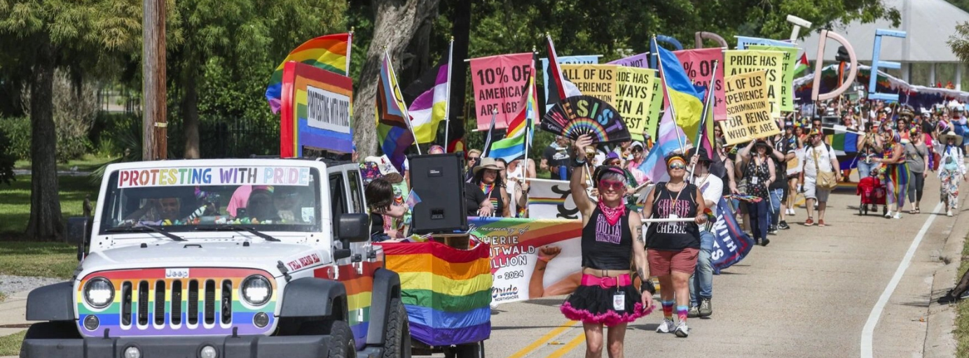 A large group of people participating in a pride parade, holding rainbow flags and banners. There is a white jeep decorated with rainbow colors and a sign that reads 'Pro testing with pride.' Some marchers are wearing rainbow-themed clothing and accessories, walking along a street lined with trees.
