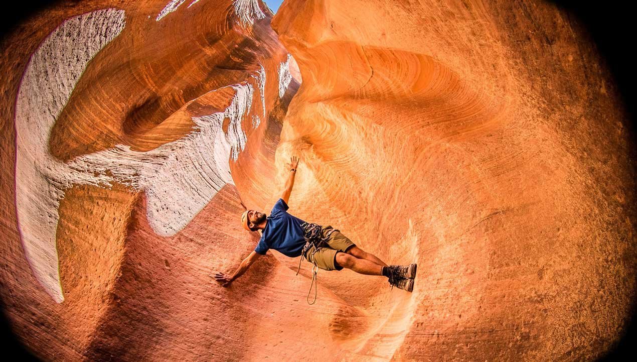 Canyoneer stems between two curving canyon walls, balancing sideways in a side plank positions, using his feet and one arm, with the other arm pointed up toward the sky.