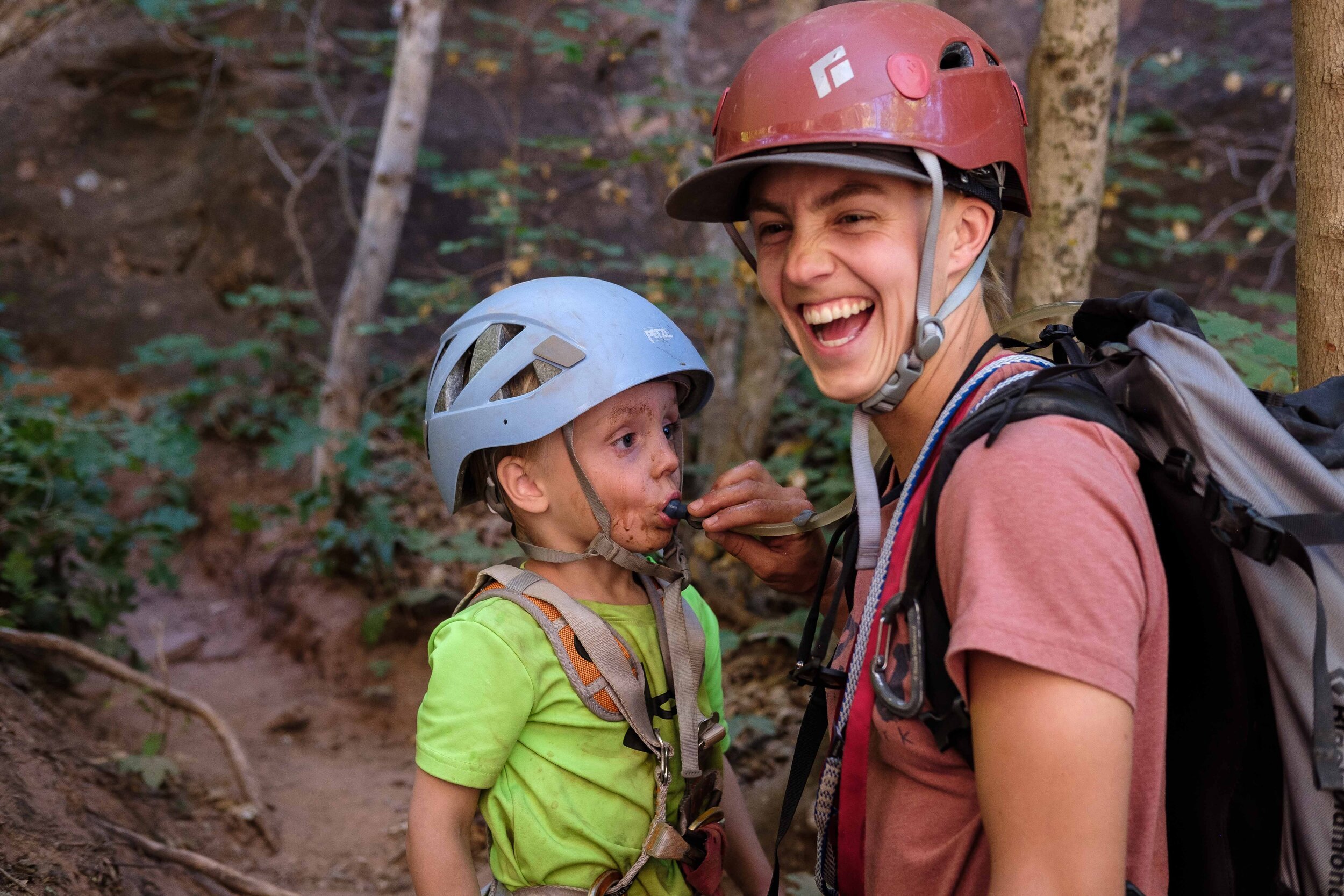 A young child drinks water from a hydration hose held by an smiling young adult on a canyon tour.