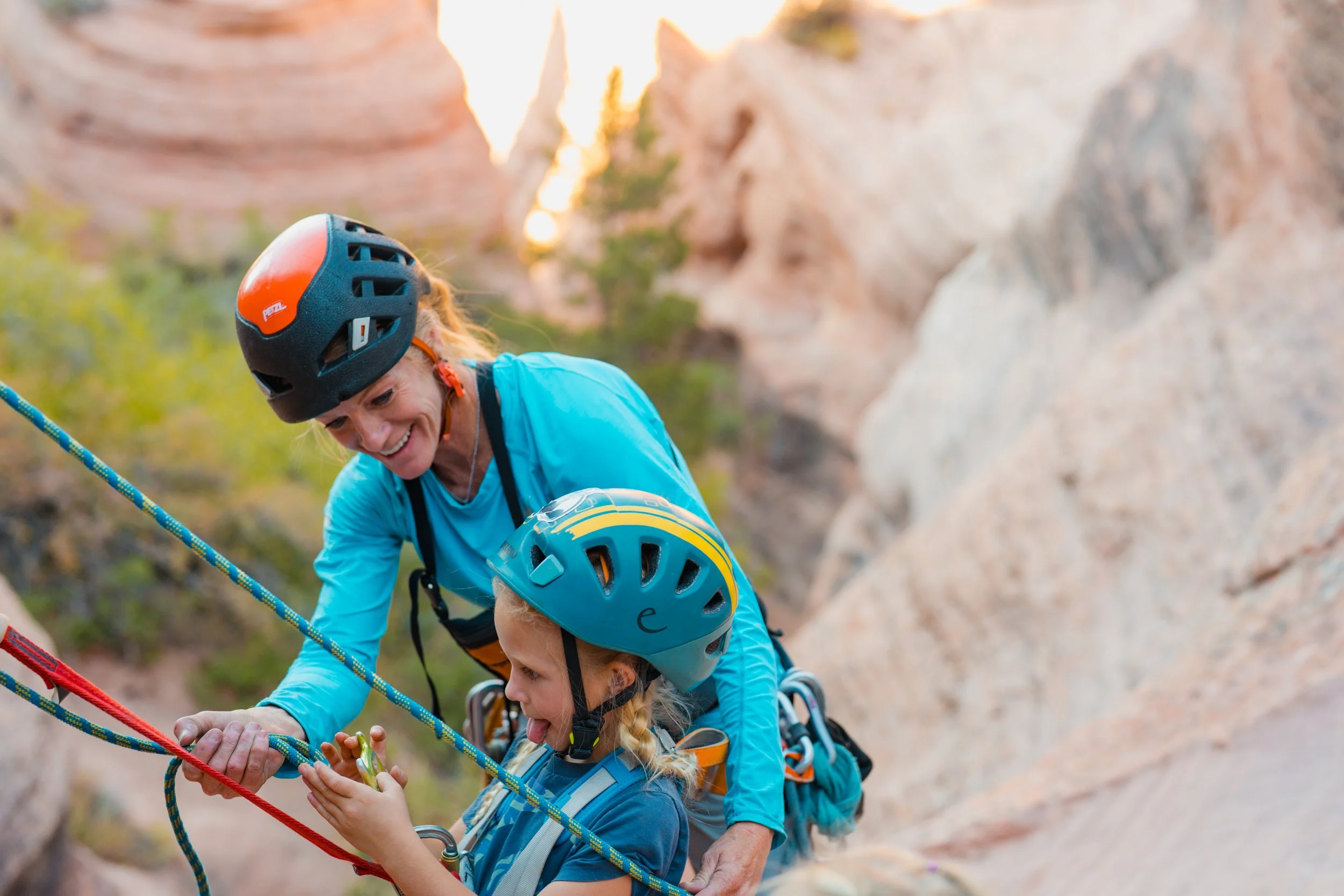 A woman and a girl, both wearing helmets and harnesses, are rock climbing together on a canyon with red rock formations, using ropes for safety and smiling.