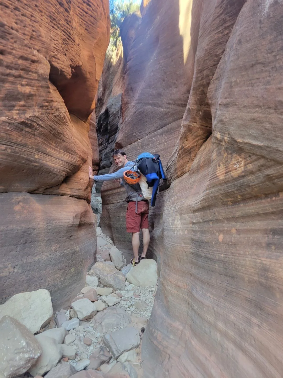 A man hiking in a narrow slot canyon with tall, smooth, reddish-brown rock walls, carrying a backpack and wearing hiking gear.
