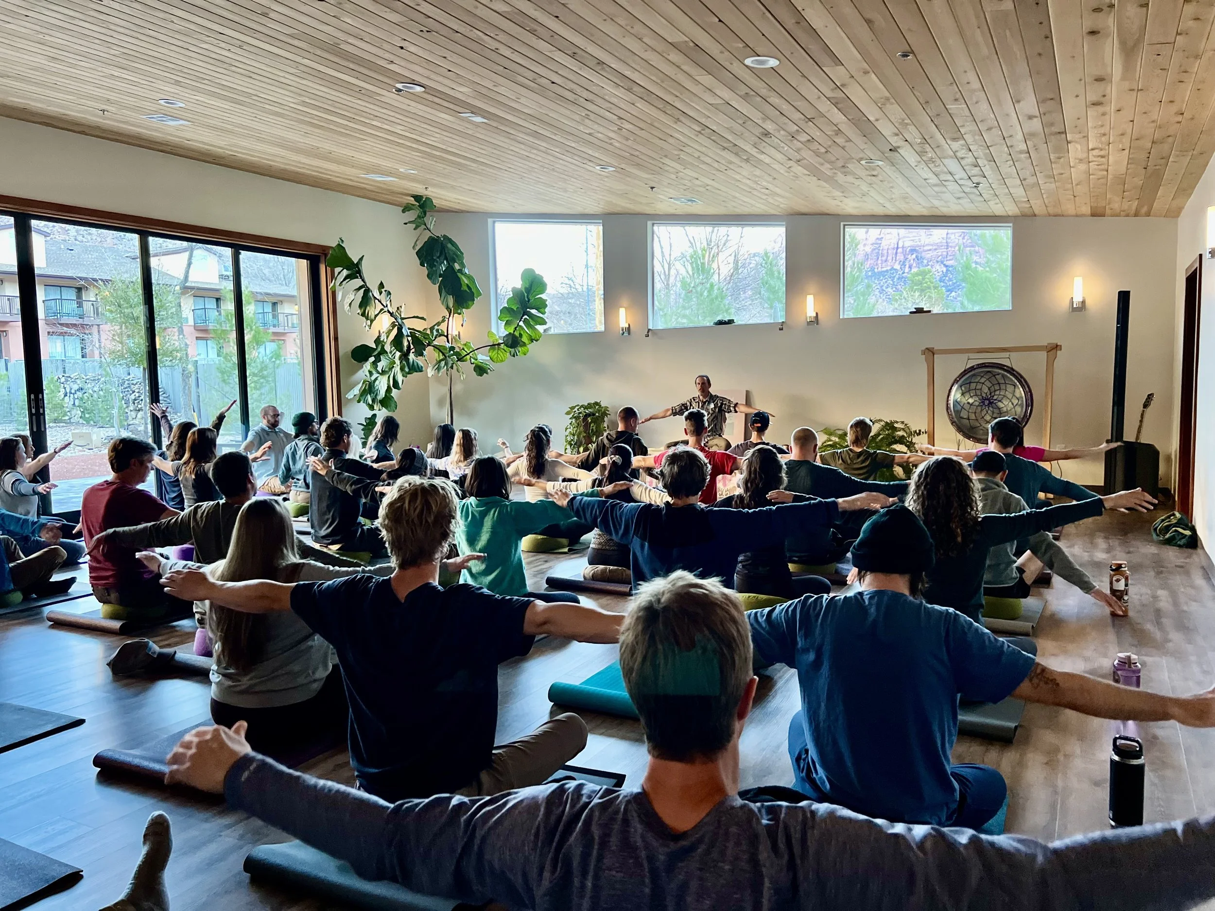 People participating in a yoga class in a spacious, sunlit room with large windows and wooden ceiling, practicing seated stretches with arms extended.