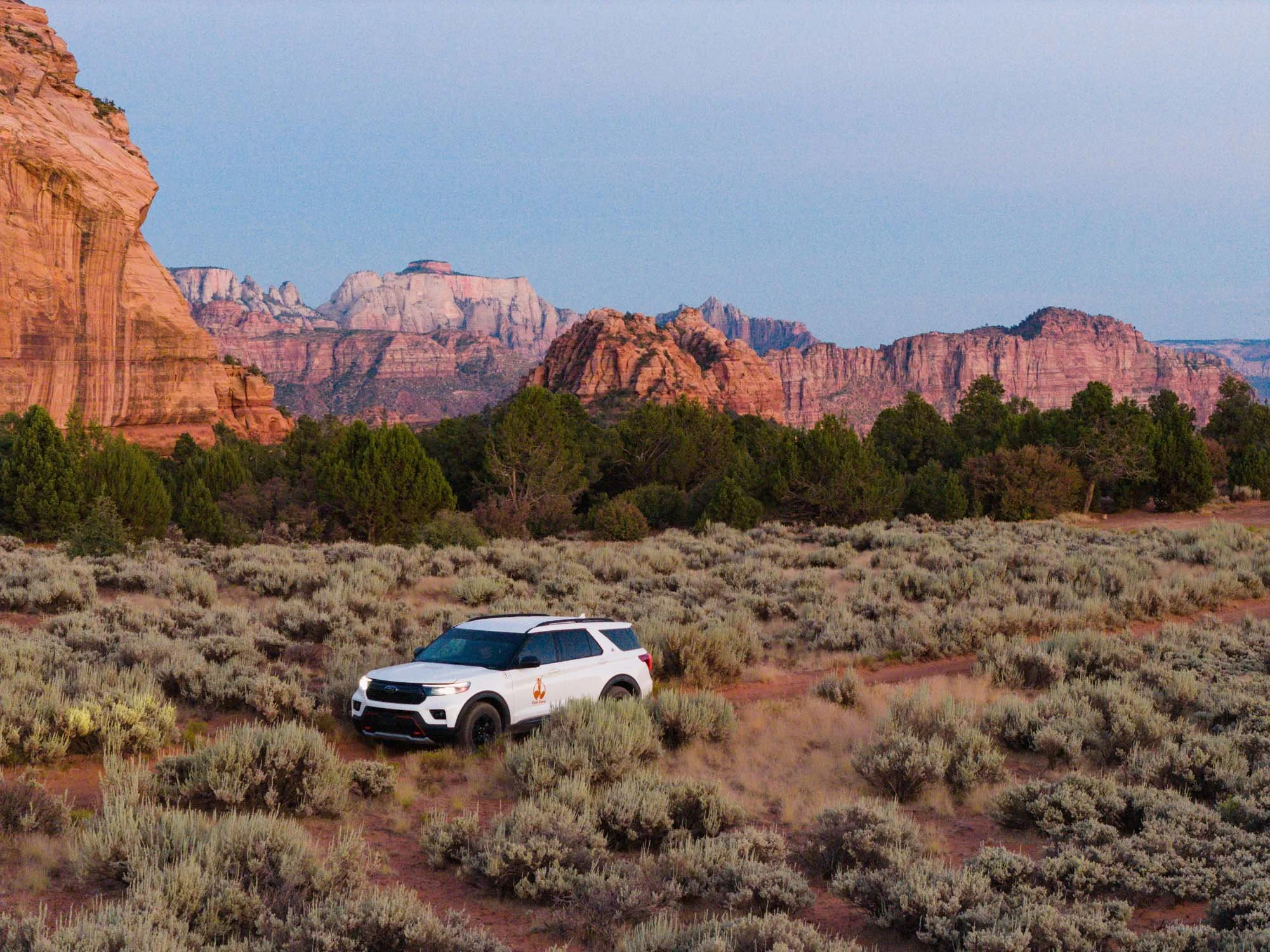 A white SUV driving through a desert landscape with low bushes and green trees, with red rock formations and cliffs in the background, under a clear blue sky.