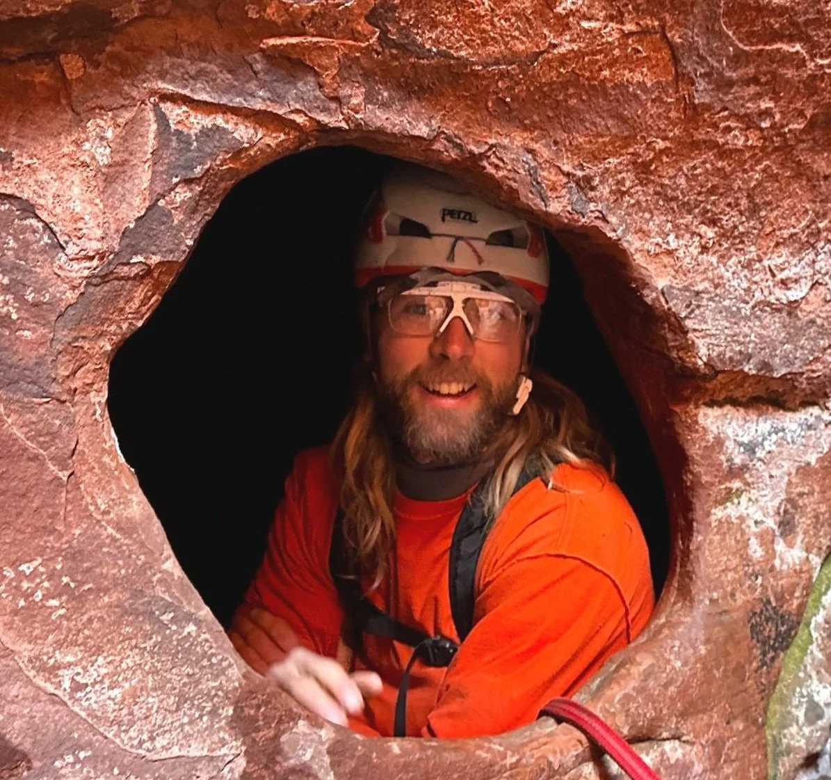 Person wearing a helmet and glasses, emerging from a small cave or rock formation with red sandstone walls.