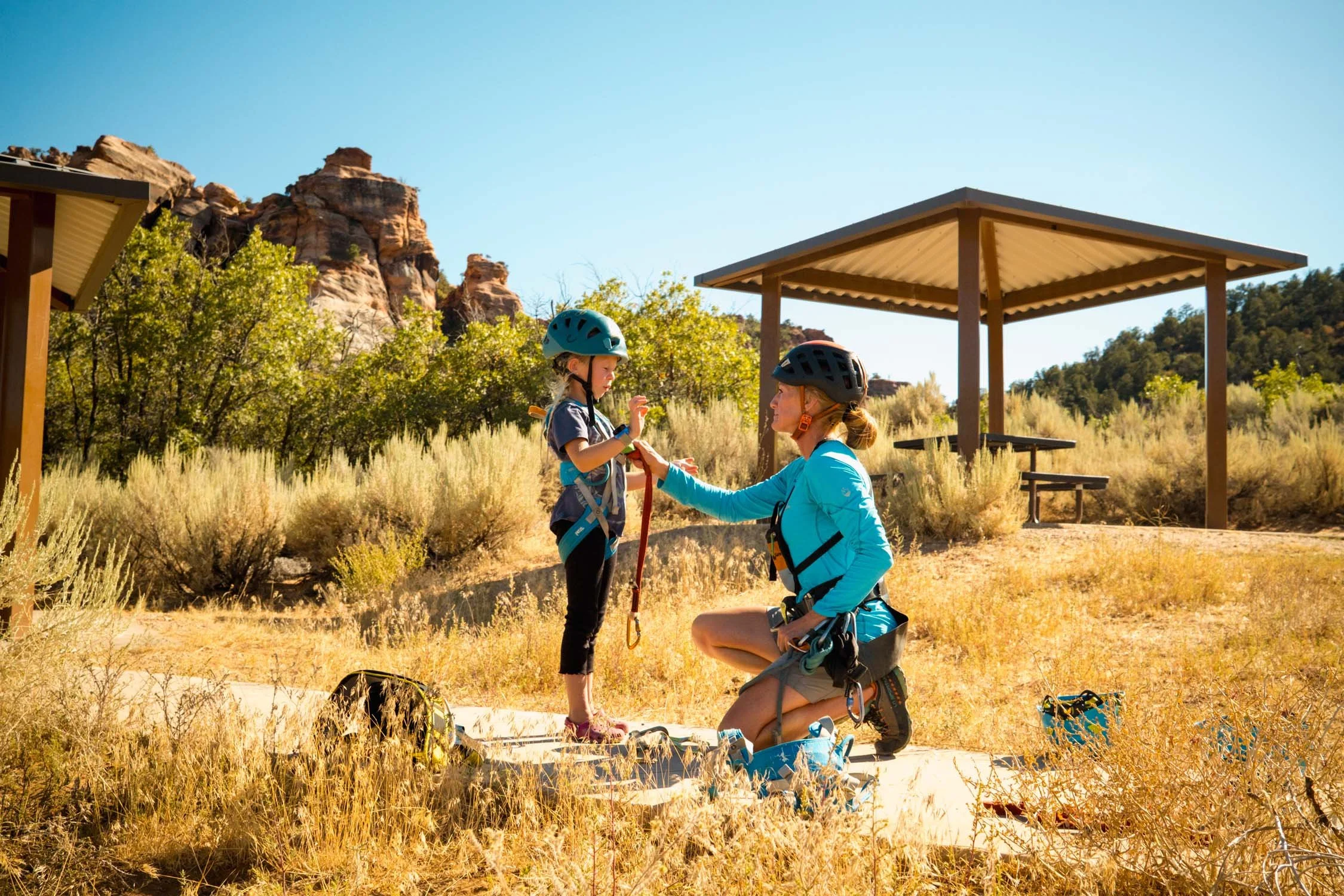 A woman kneeling and helping a young girl with zip-lining gear outdoors on a sunny day with trees, shrubs, and rock formations in the background.