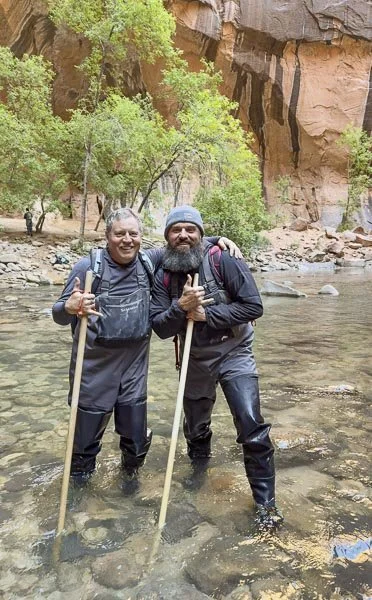 Two men wearing waders and holding walking sticks stand in a shallow river with a rocky and tree-lined background.