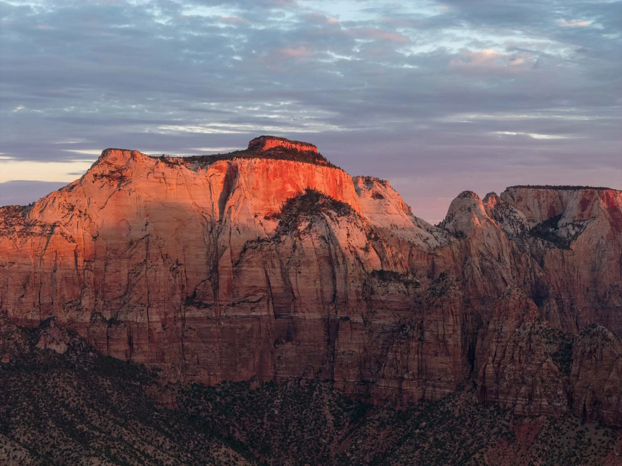 Sunset over Zion National Park with towering, red-orange rock formations and a partly cloudy sky.