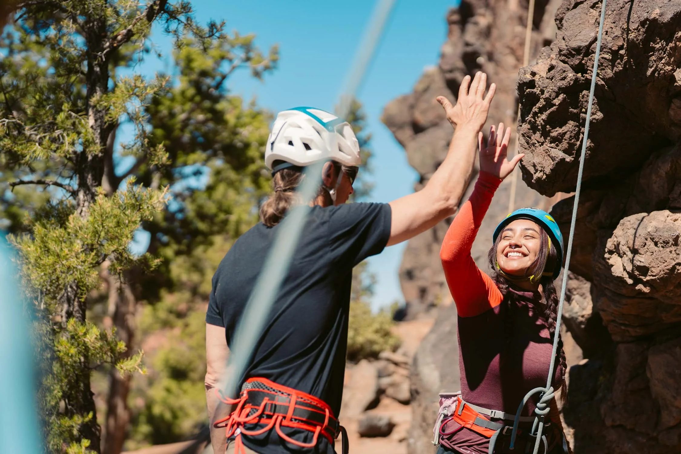 A woman and a man high-fiving while rock climbing outdoors, both wearing helmets and harnesses.
