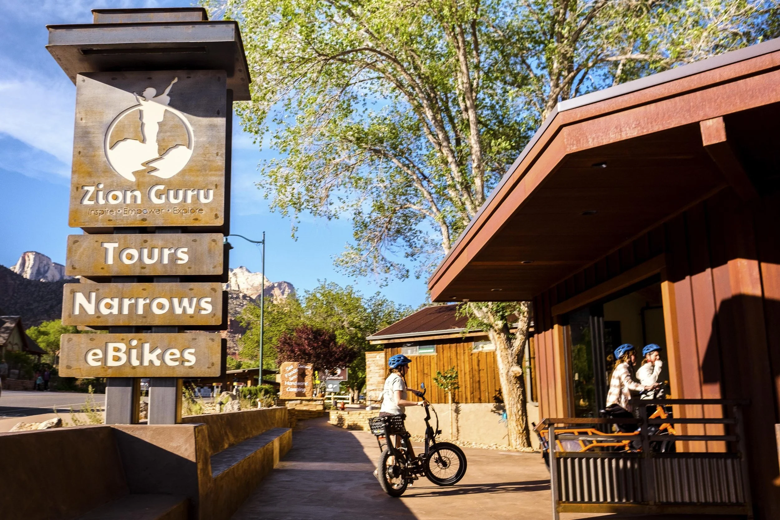 Zion Guru sign advertising tours, Narrows hikes, and eBikes; people in helmets with bikes; trees and buildings in the background.