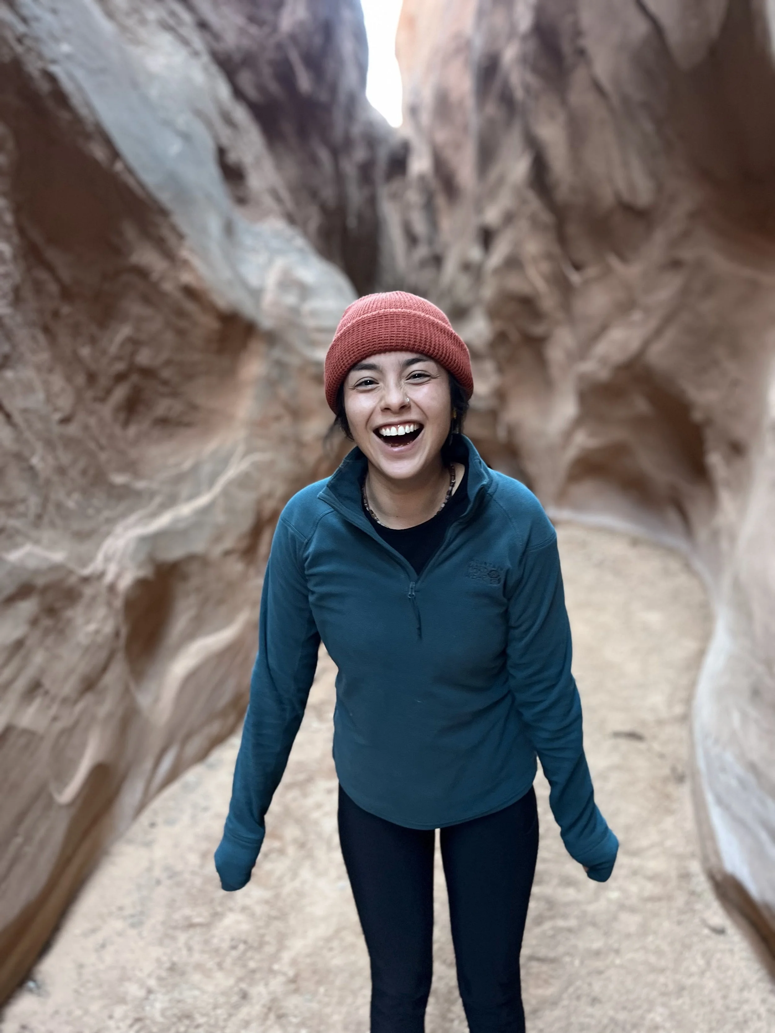 Person wearing a beanie and sweater standing in a narrow rock canyon, smiling.