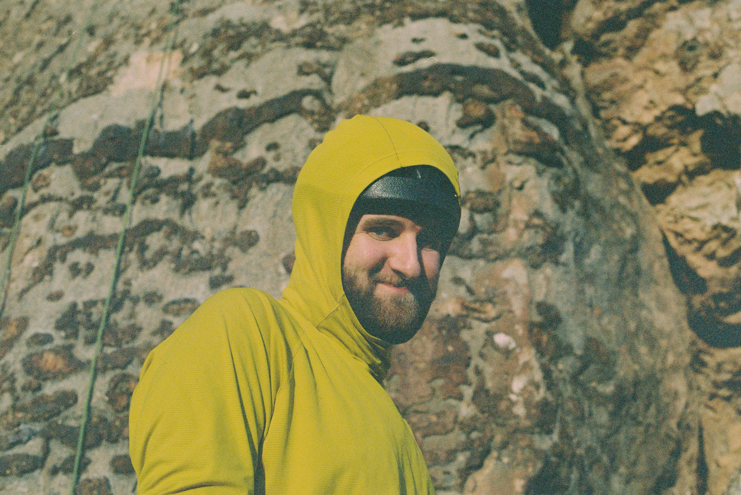 A man in a yellow hooded jacket and black helmet standing in front of a rocky, textured wall.