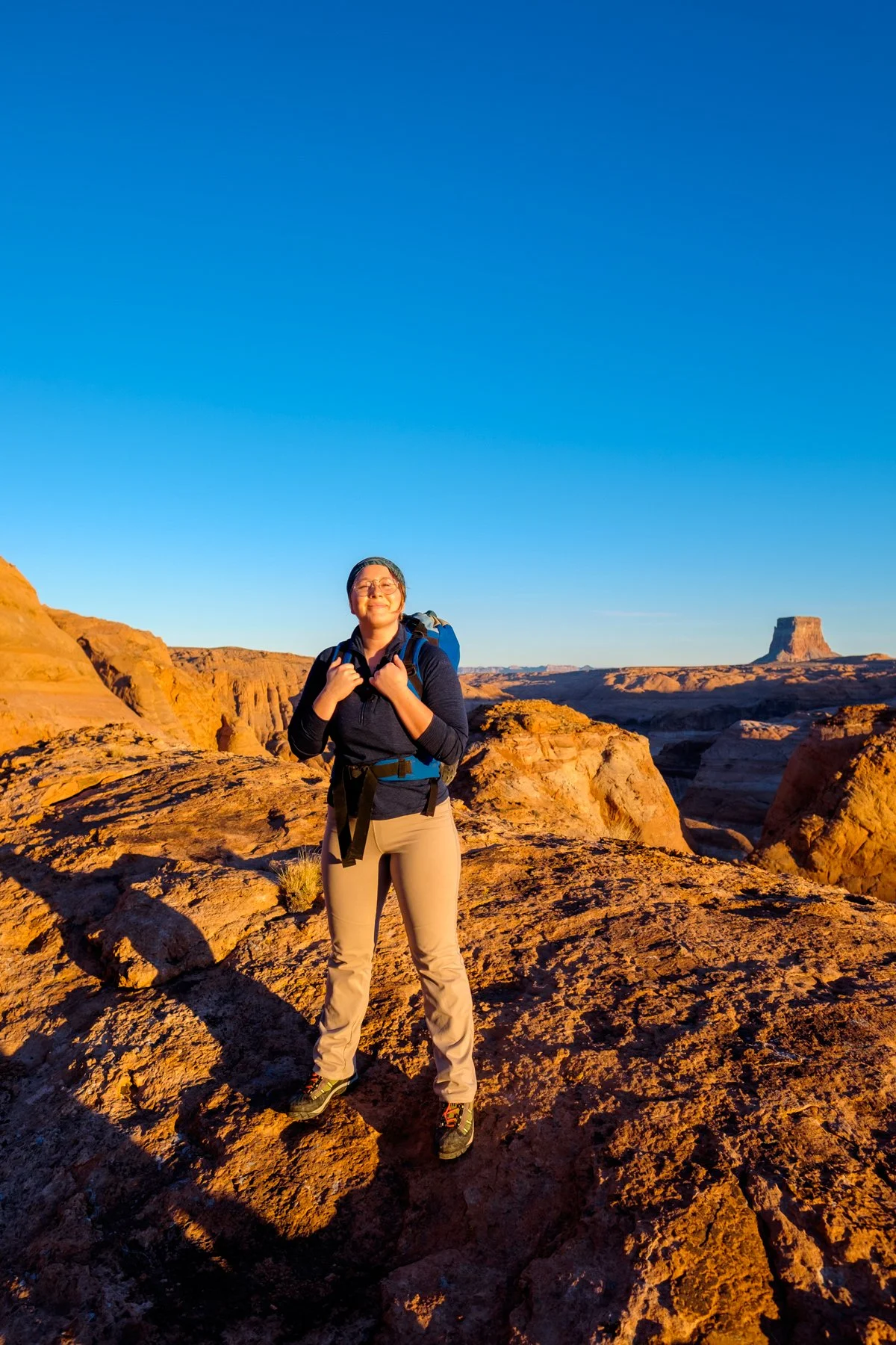 A woman standing on a rocky desert landscape, smiling, wearing hiking gear and carrying a backpack, during sunset with sparse vegetation and distant rock formations.