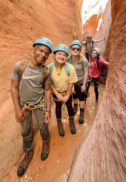 Group of people canyoning in a narrow, sandy slot canyon wearing helmets and backpacks.