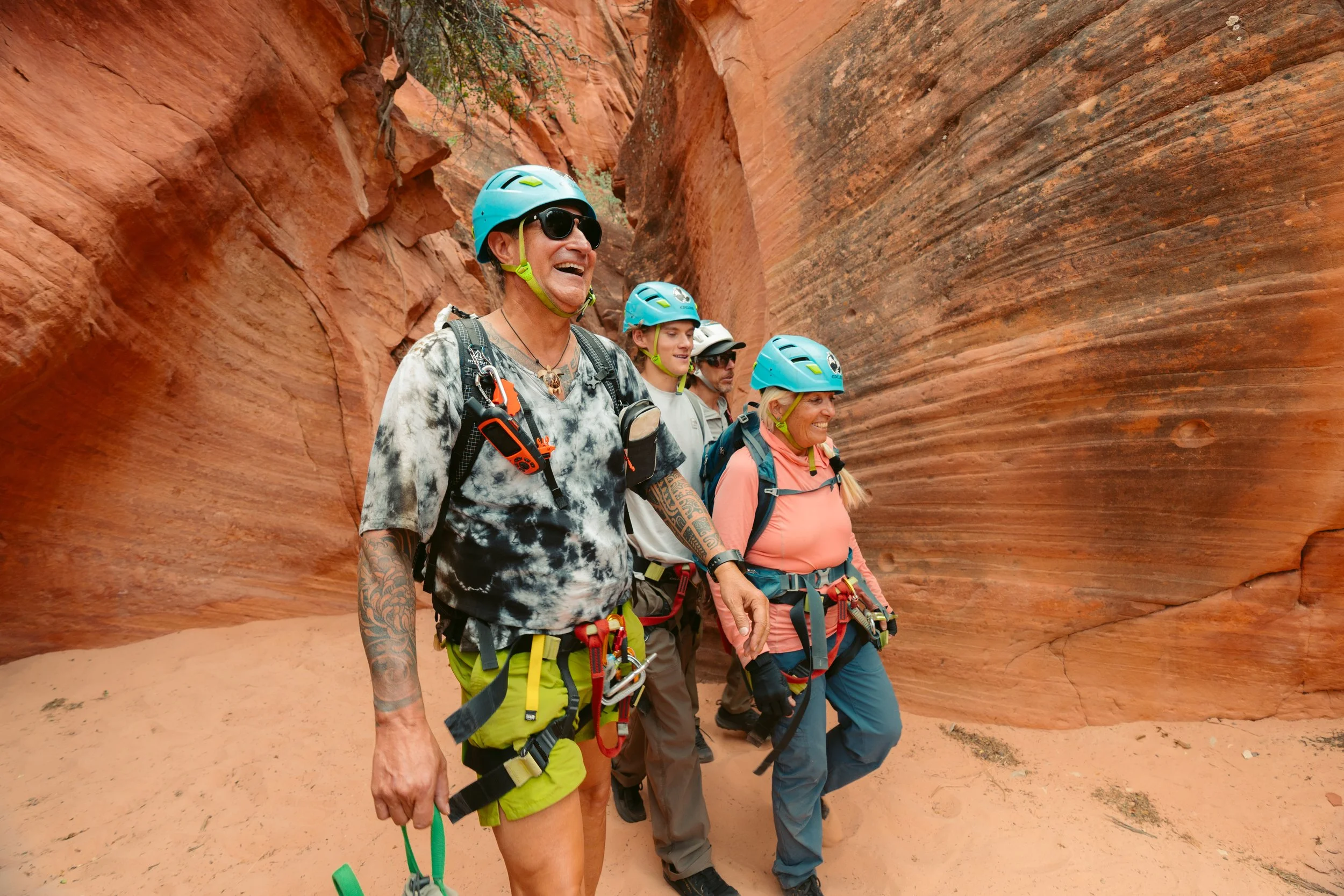 Three smiling hikers walking on a trail in a mountainous desert landscape with trees and red-rock formations in the background.