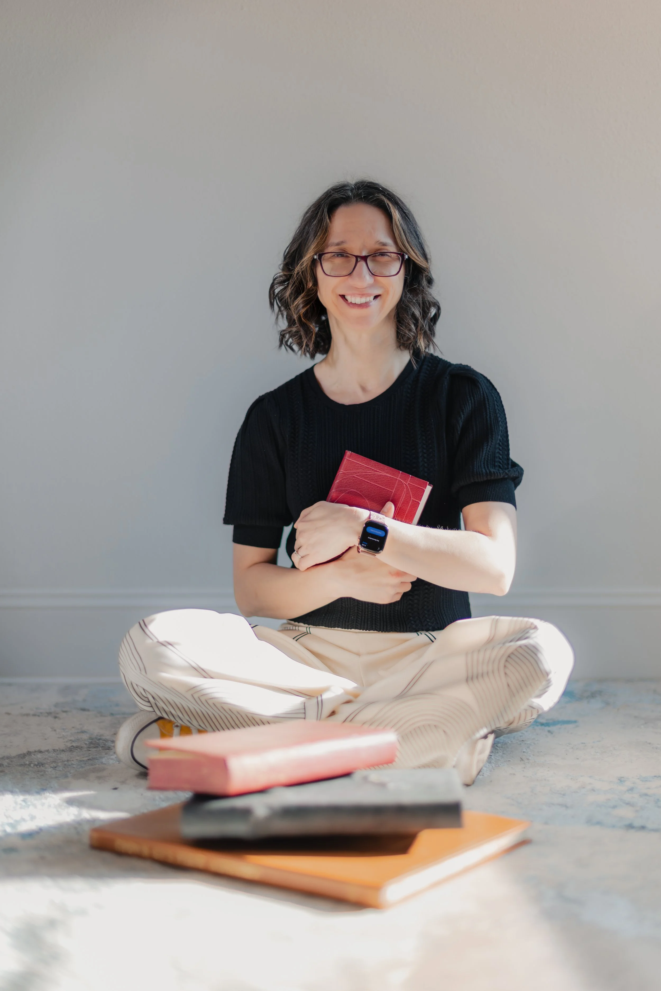 Woman sitting cross-legged on a carpeted floor, holding a red book, wearing glasses, a black shirt, and beige pants with thin stripes, with a smartwatch on her wrist, smiling at the camera with books around her.