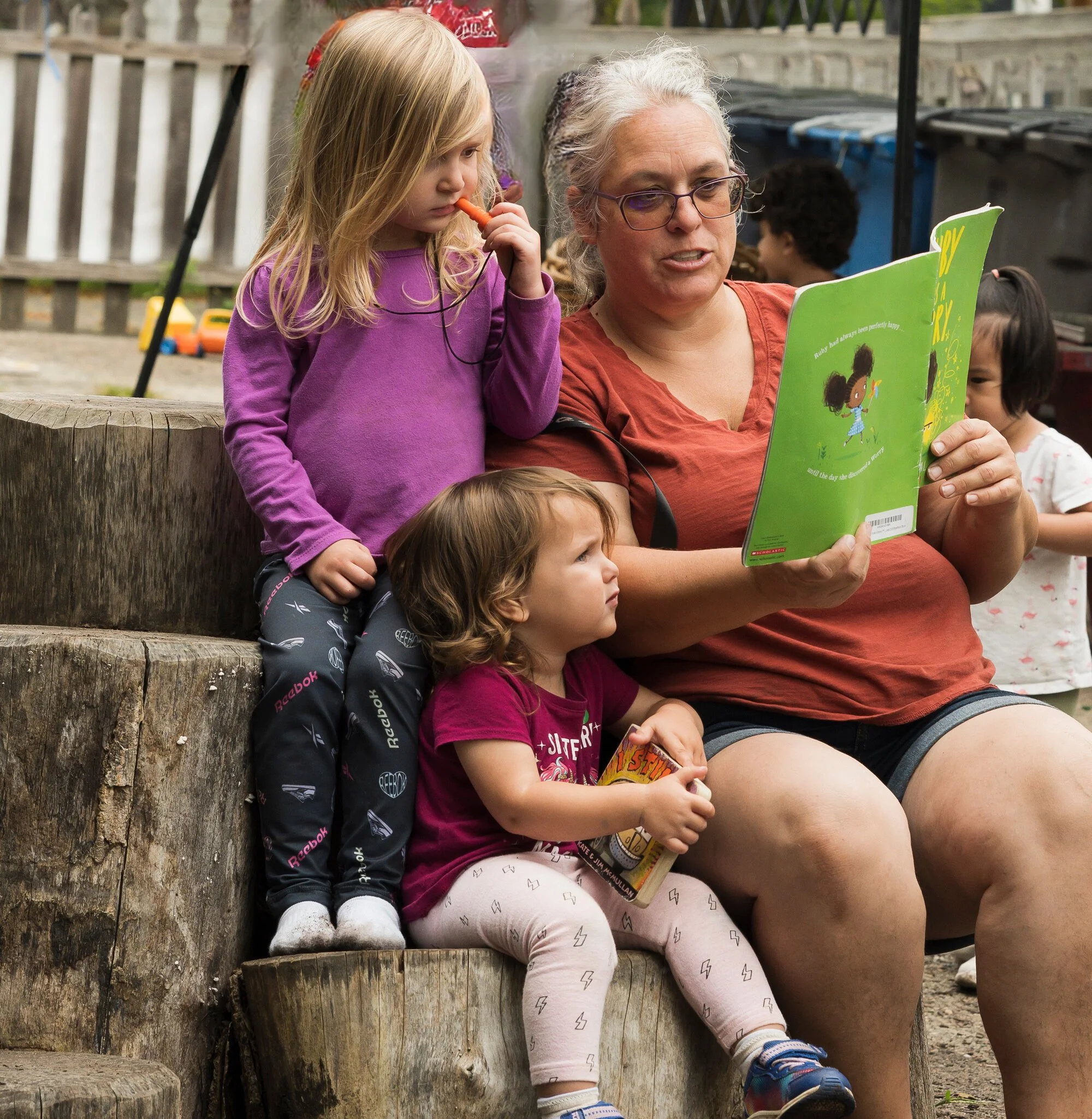 Two children, one with blonde hair and one with light brown hair, both light skin tone, sit on stumps and are actively engaged with the book, "Ruby Finds a Worry", that a teacher with glasses and light skin tone is reading.