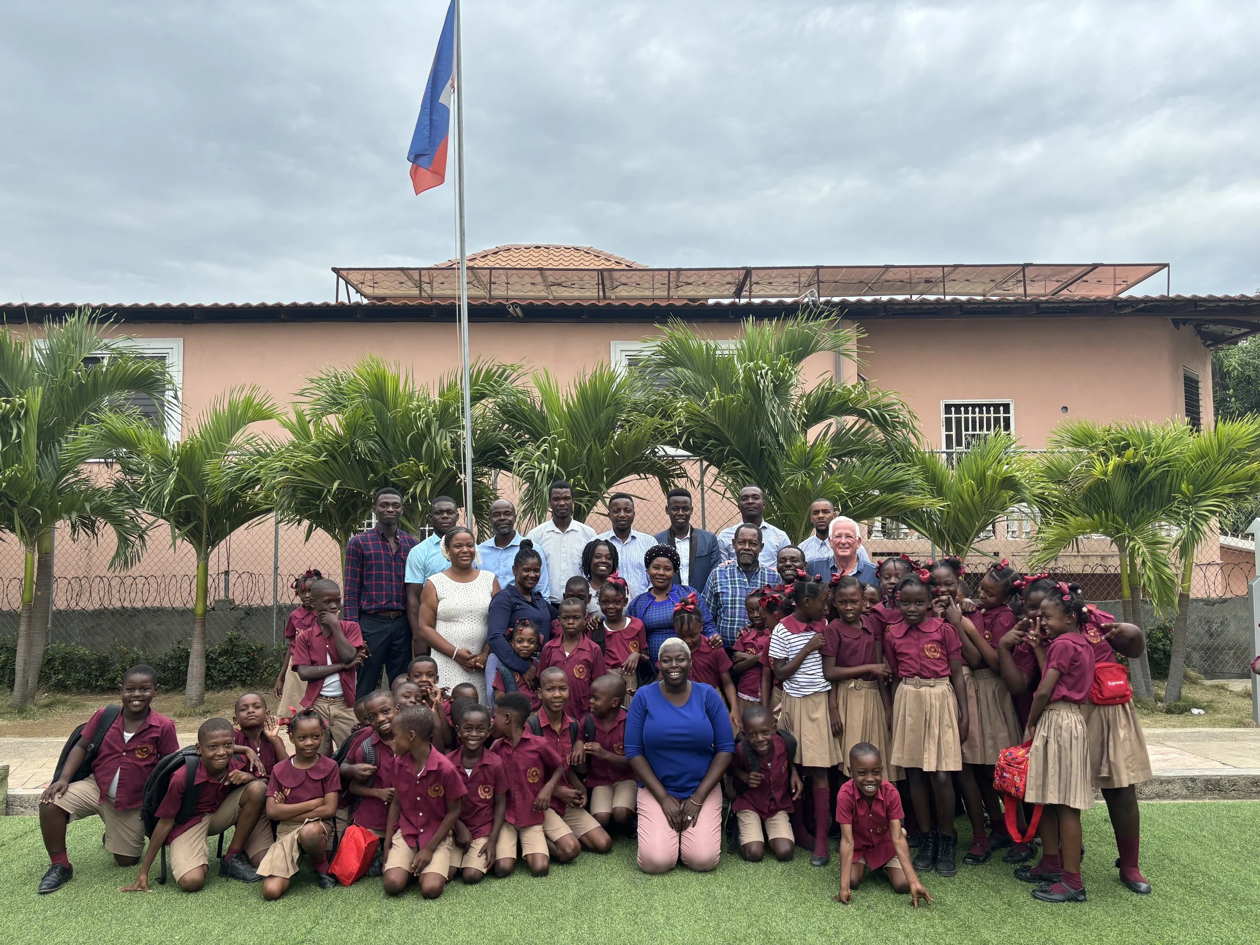 A large group of school children and several adults posing for a photo outside in front of a pink building with a flagpole. The children are wearing maroon uniforms and standing among trees and greenery.