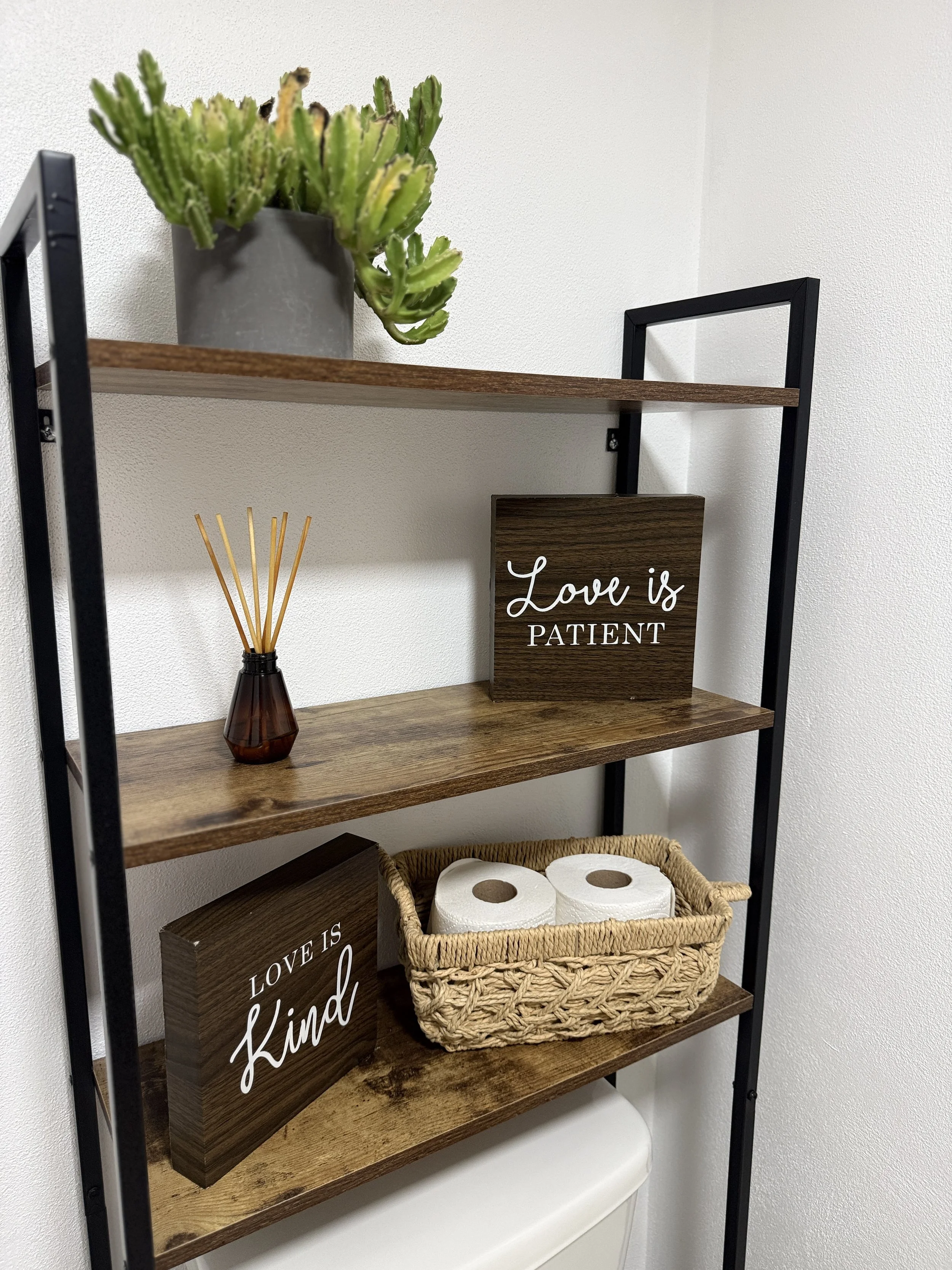 A shelving unit with a potted cactus on top, a reed diffuser, two decorative wooden signs that read "Love is Patient" and "Love is Kind," and a basket with two rolls of toilet paper.