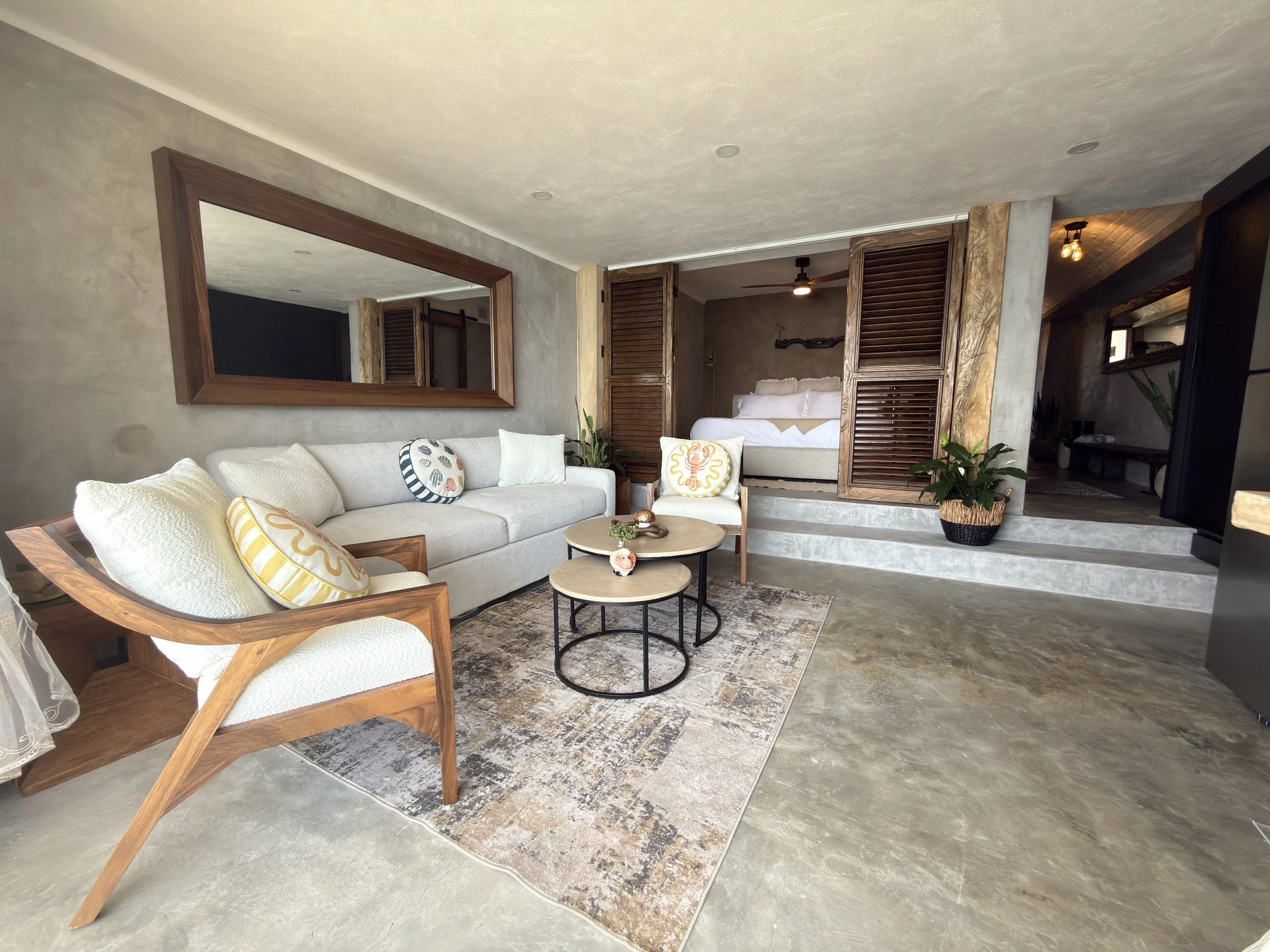 Living room with a beige sofa, wooden armchair, round coffee tables, colorful pillows, large wall mirror, and a bedroom with a bed enclosed by wooden sliding doors in the background.