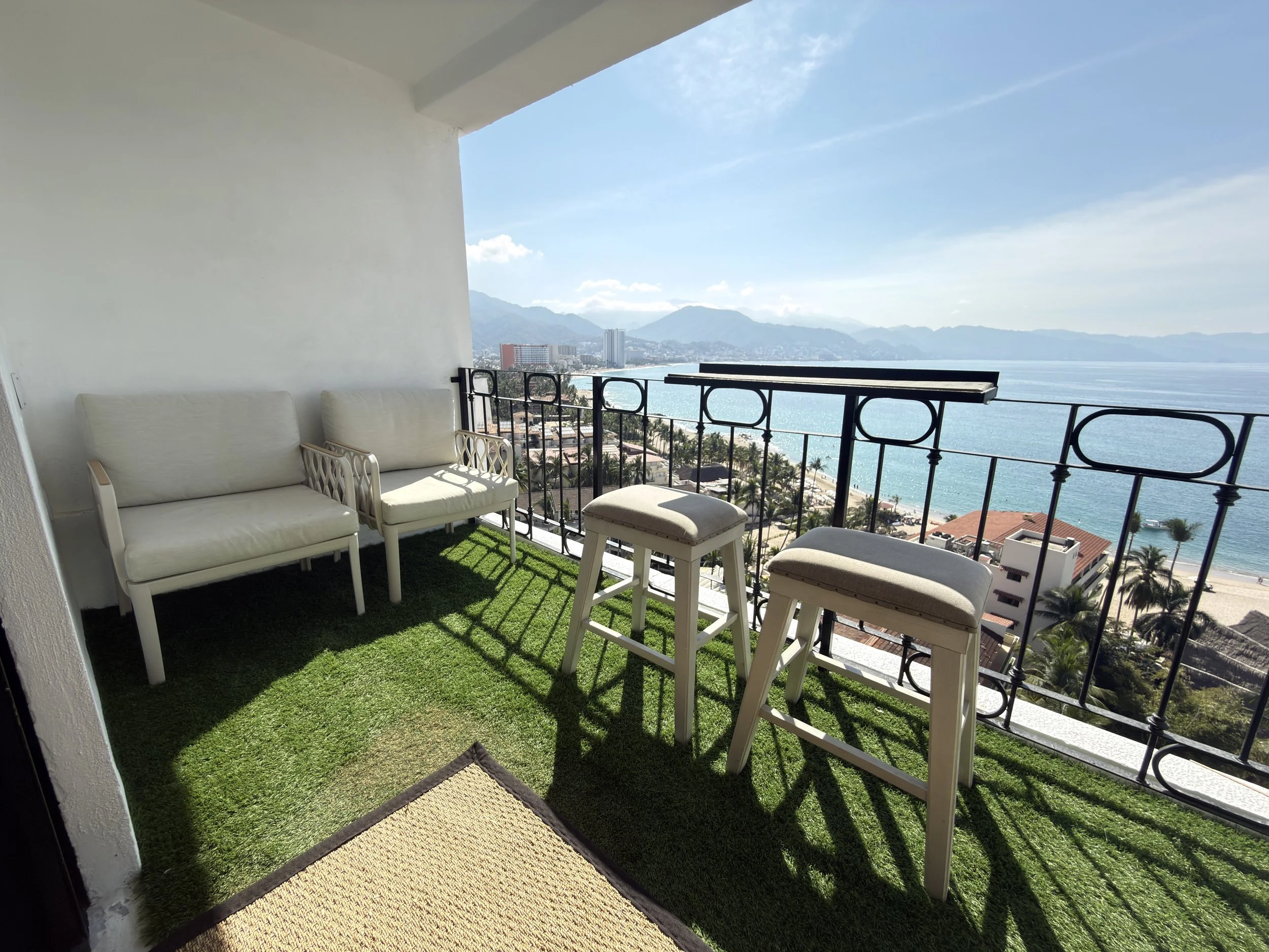 Balcony with white outdoor furniture, two chairs, and two stools, overlooking a beach with clear water, palm trees, and mountains in the distance.