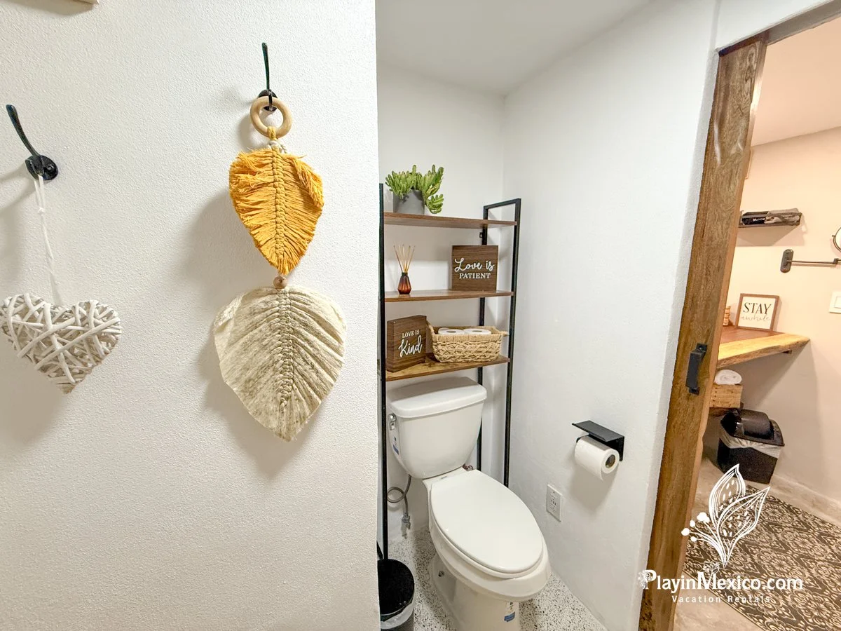 A bathroom with a toilet, decorative wall hanging of hanging leaves, wooden signs that read "Love is PATIENT" and "Love is Kind," and a shelf with a plant and baskets. The bathroom door is wooden, and there is a toilet paper holder with toilet paper.